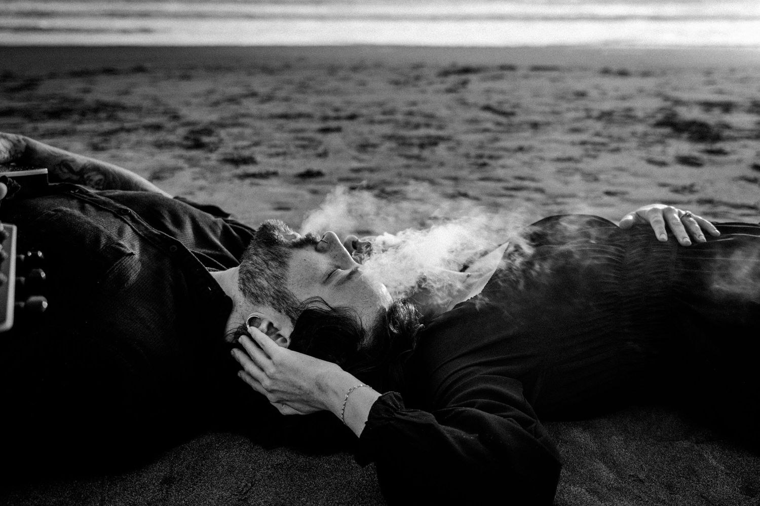 Black and white photo of a couple kissing on a beach, smoke swirling around them, with a guitar nearby.Photography by The Y.K. Studio.