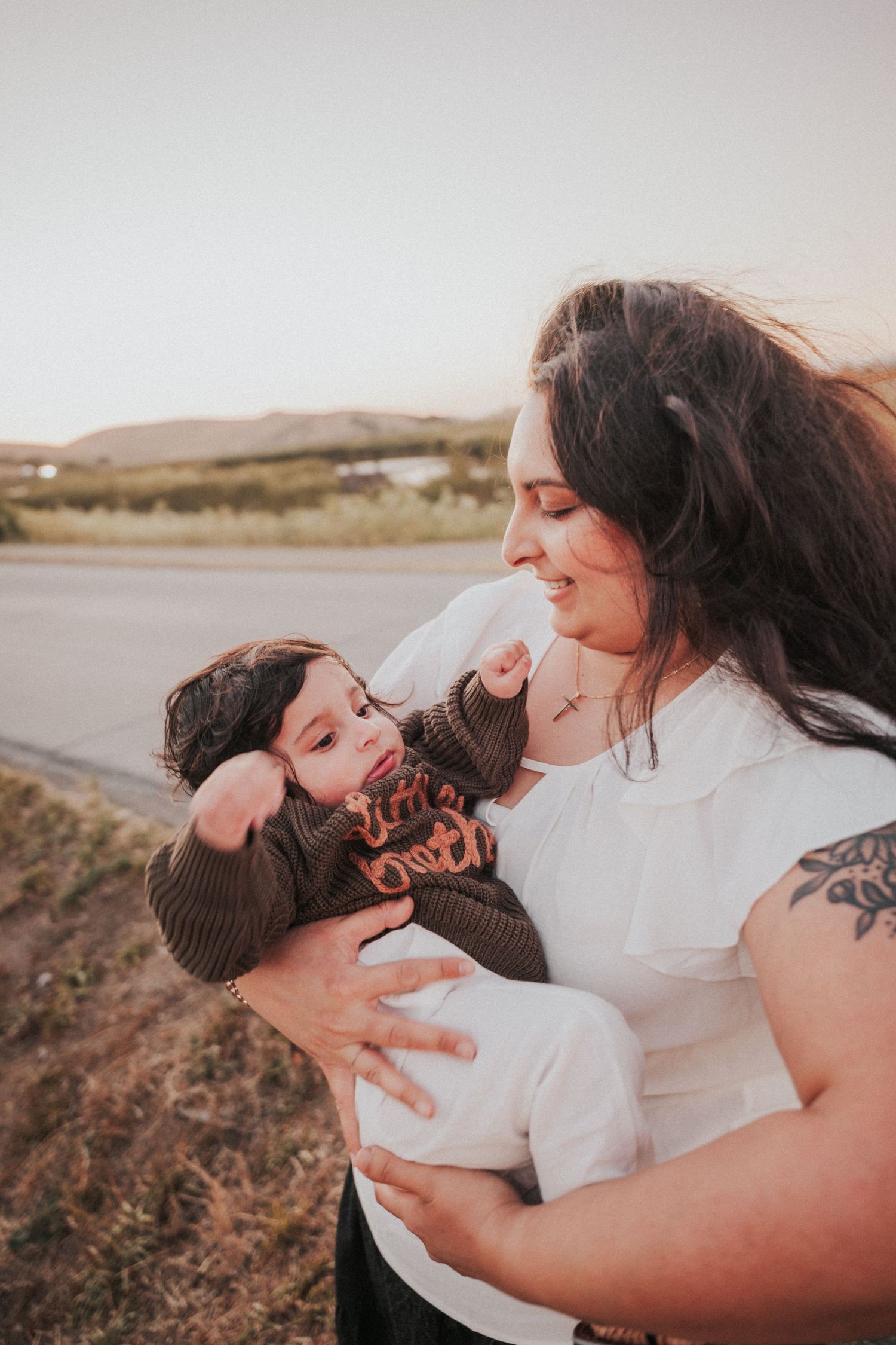 A woman with long dark hair and tattoos lovingly holds a baby outdoors near a road at golden hour.