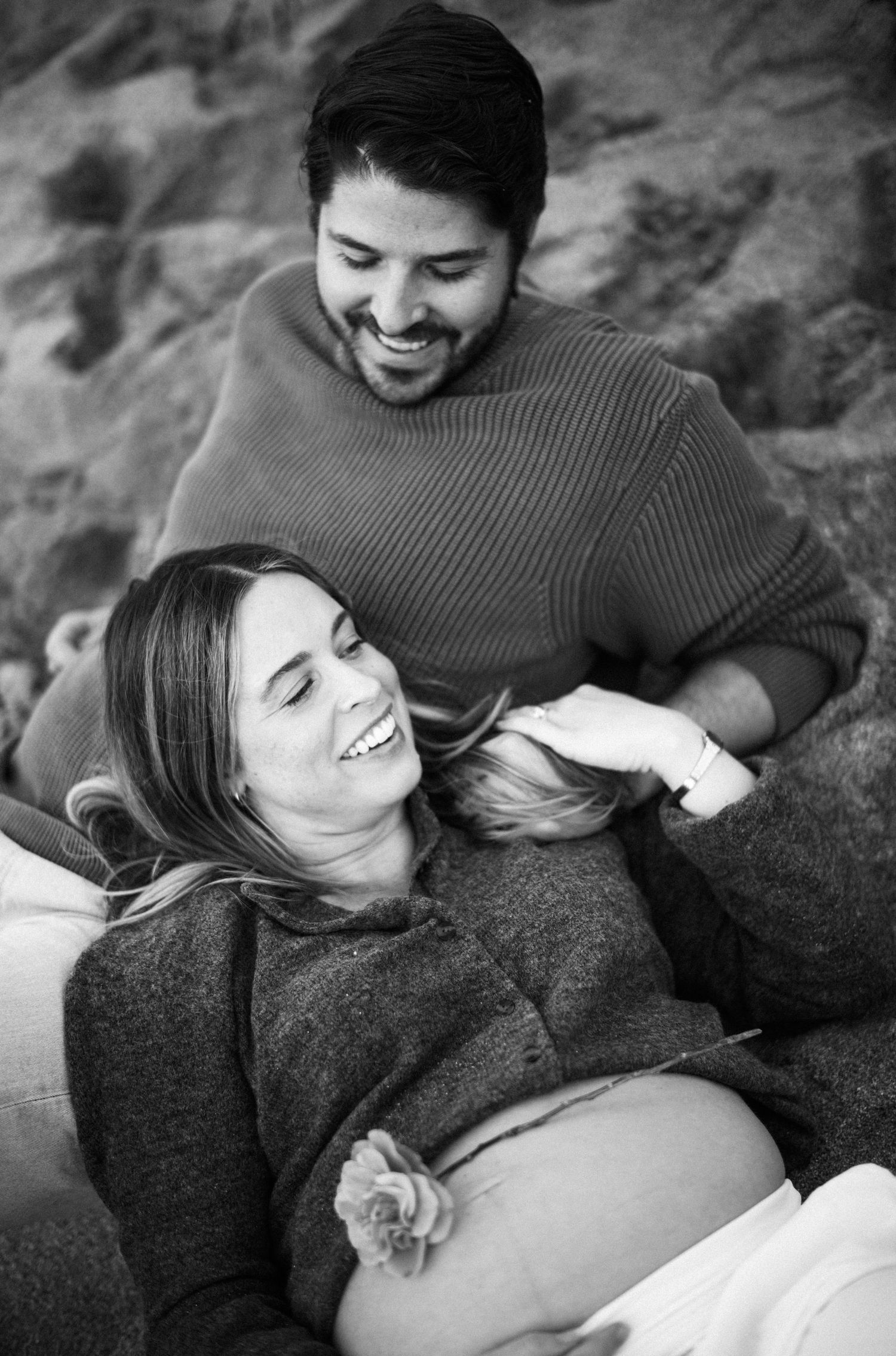 Black and white maternity photo of a smiling pregnant woman reclining against a laughing man outdoors.