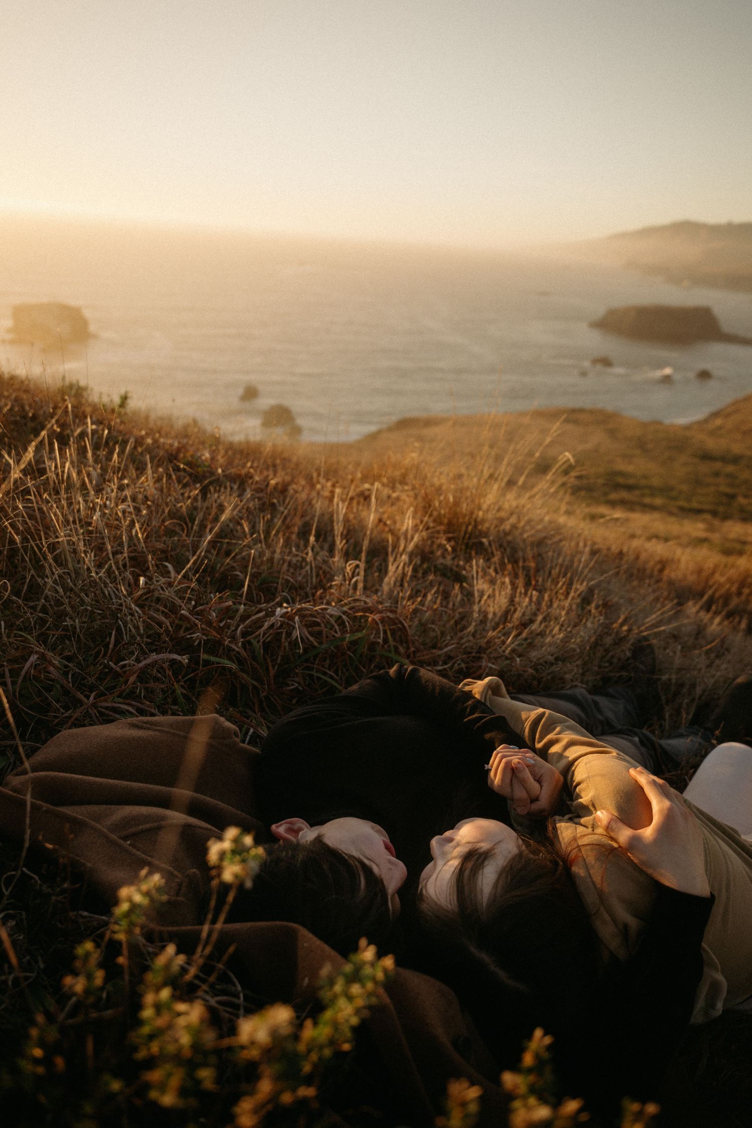 Couple lying close together in tall golden grass on a seaside bluff at sunset, with ocean rocks and hazy coastline in the distance.