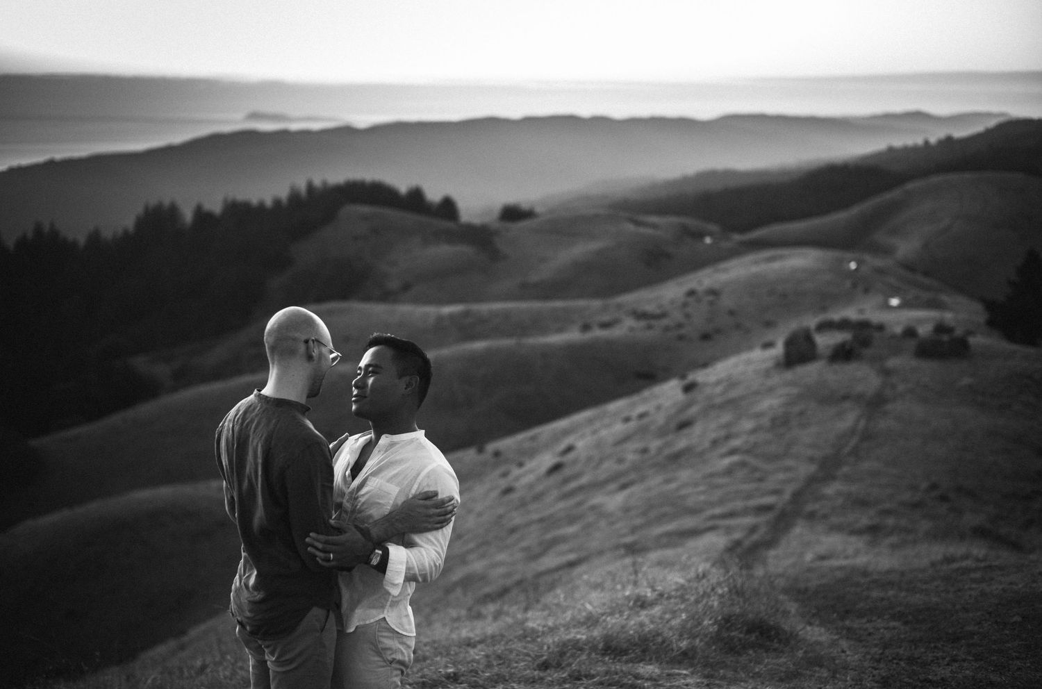 A couple embraces romantically on a scenic hillside in a black and white photo.