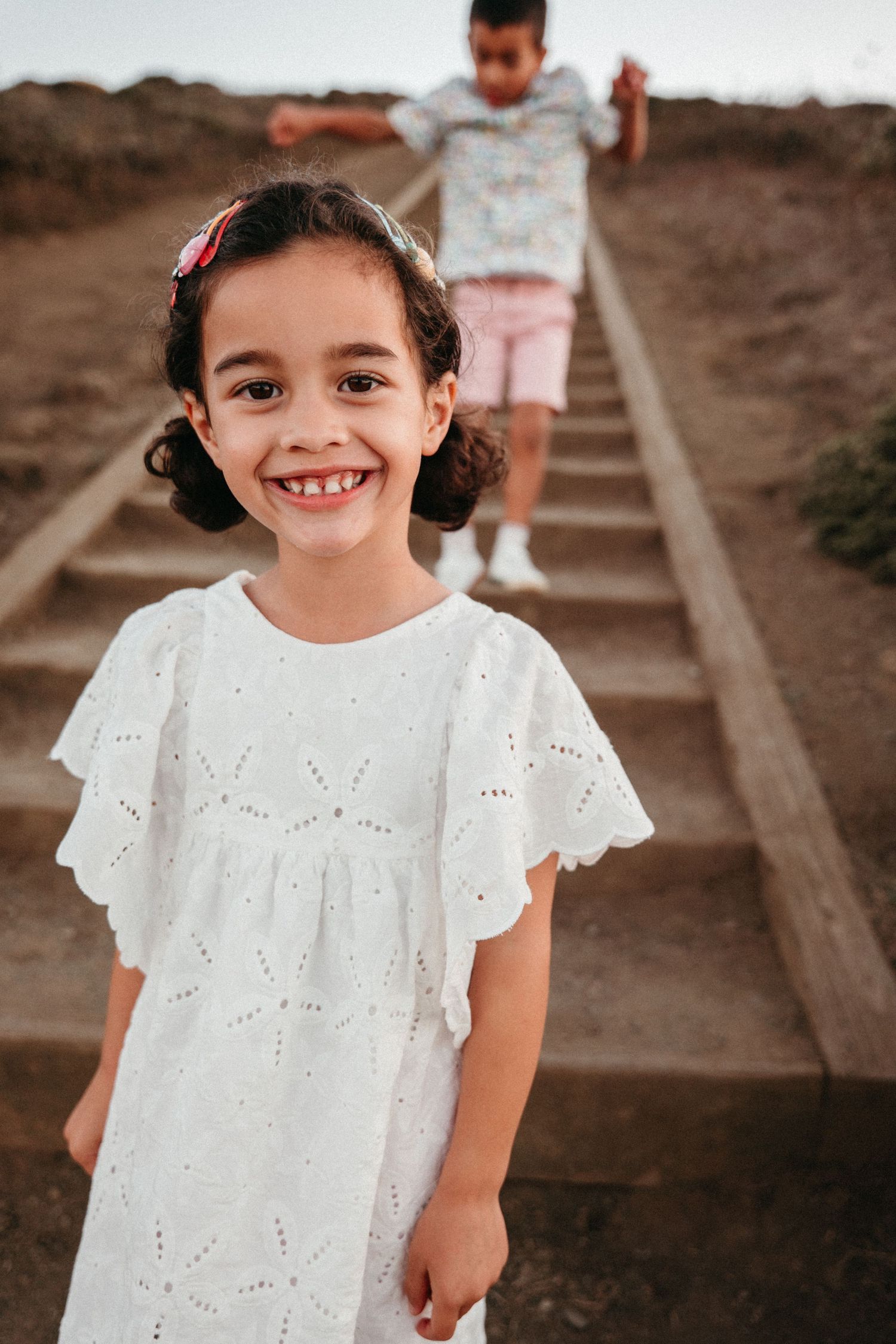 A smiling young girl in a white eyelet dress stands at the bottom of outdoor stairs, with a boy descending behind her.