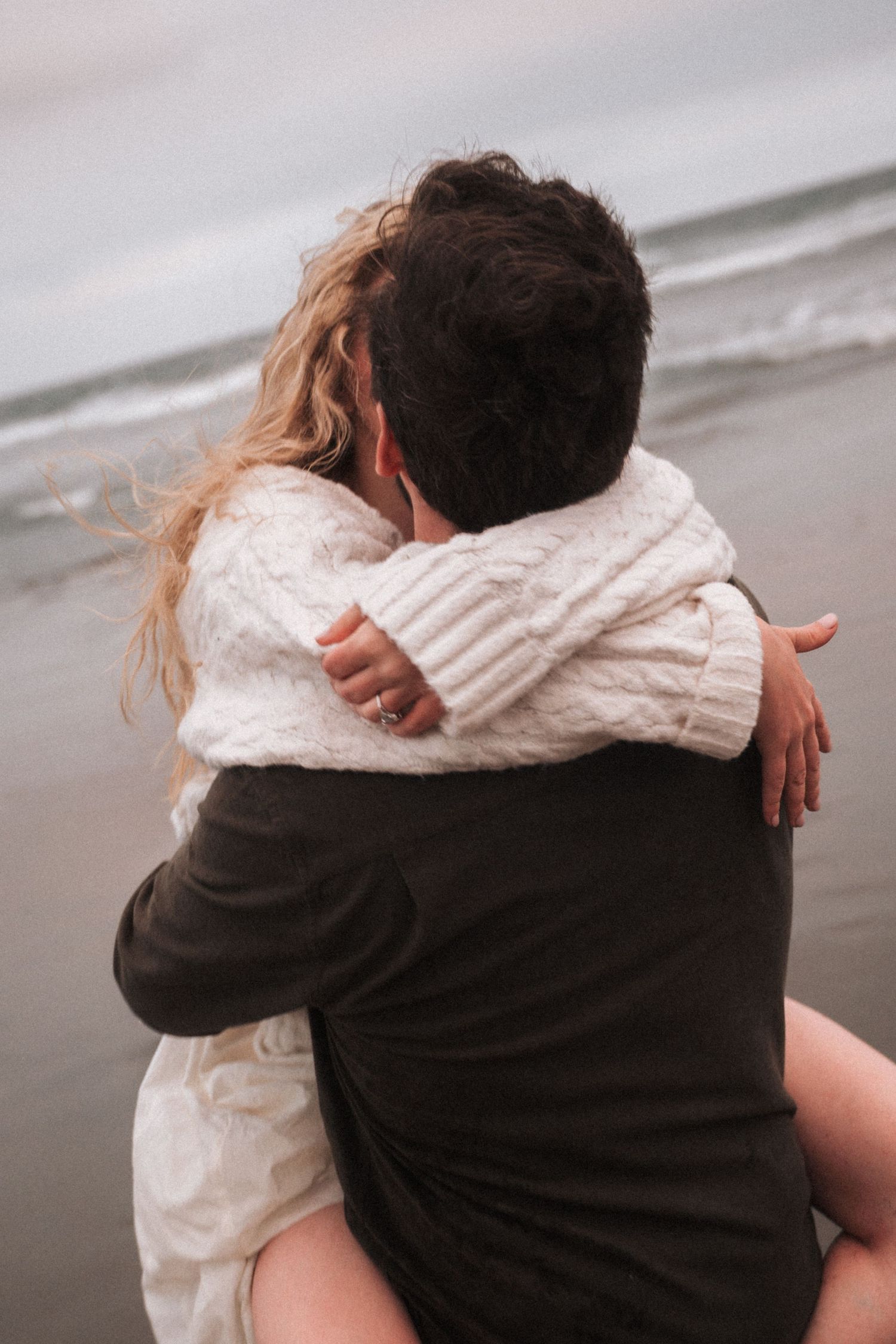 Couple embracing on a misty beach, woman in white knit sweater hugging man in black, romantic ocean backdrop.
