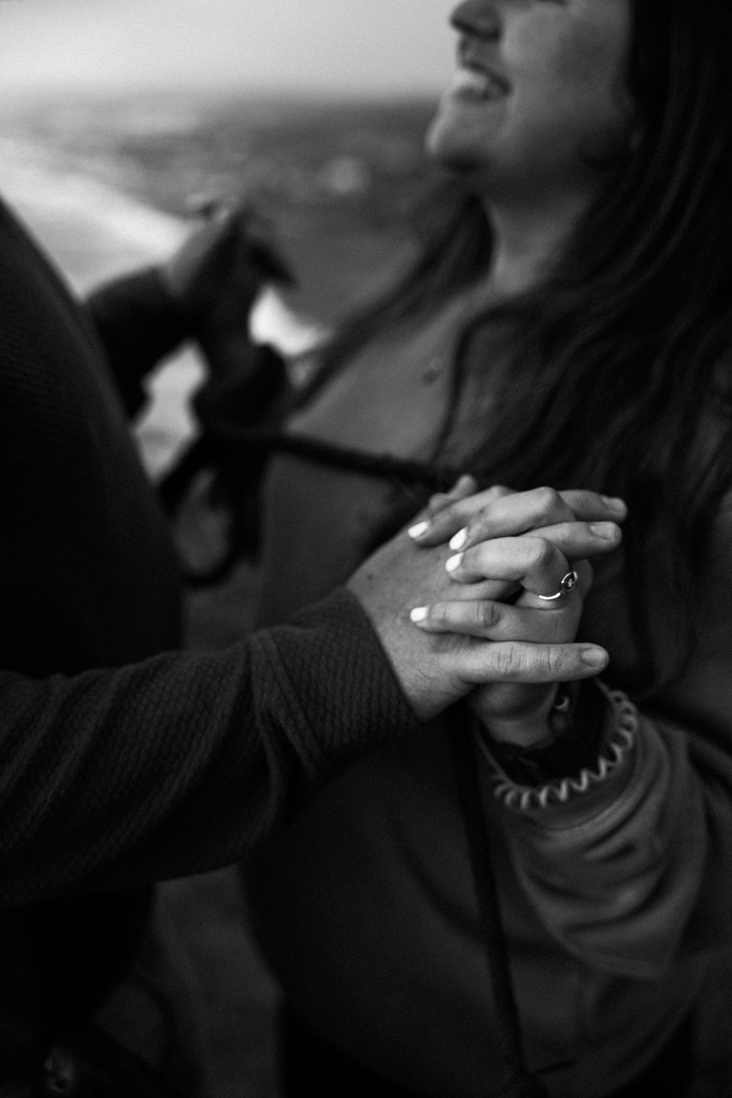 Black and white photo of couple holding hands outdoors, woman smiling, wearing a ring, romantic moment captured.