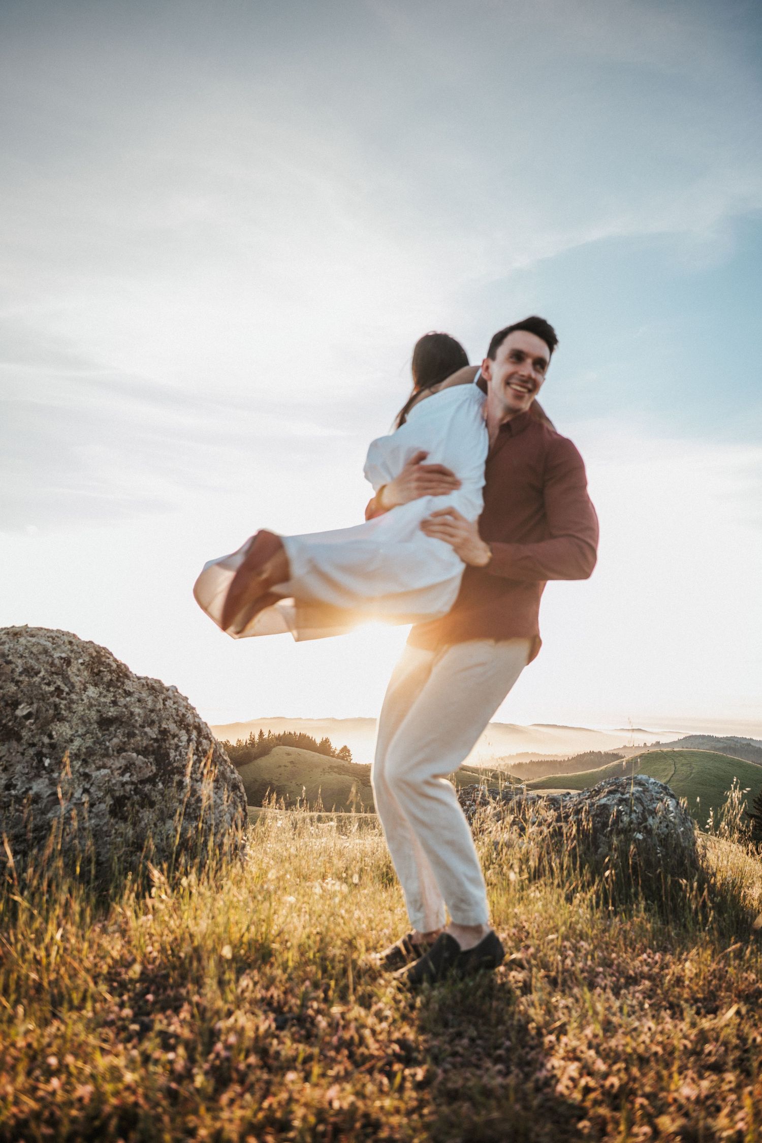 Man in red shirt joyfully lifting woman in white dress during golden sunset in scenic hillside field.