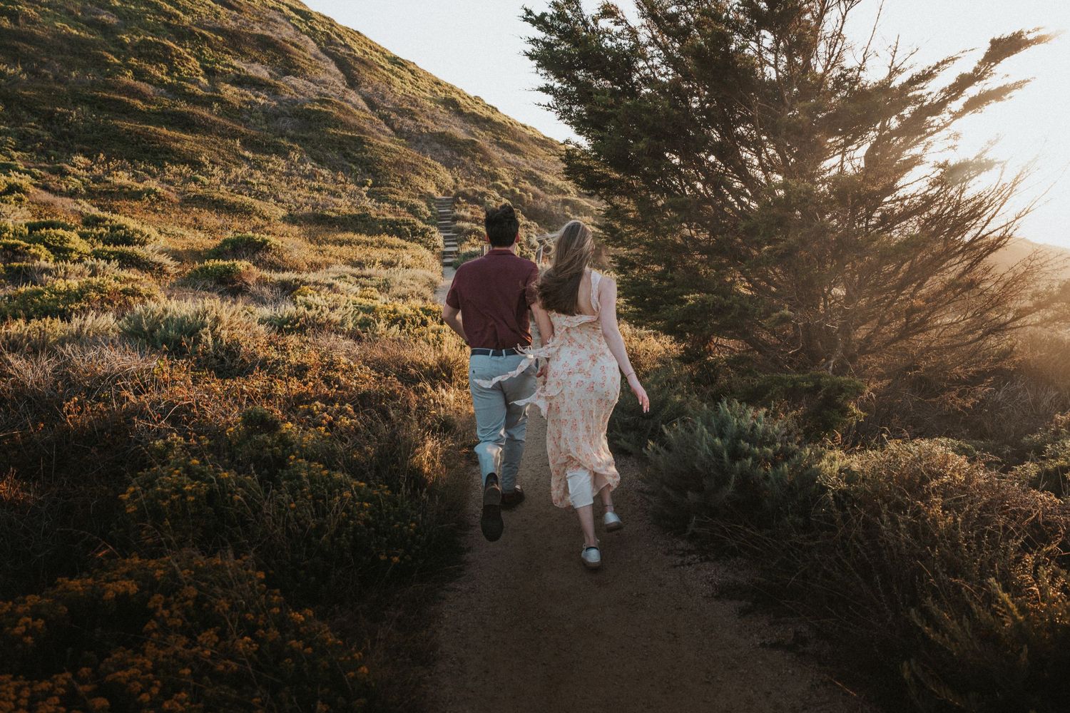 Couple holding hands running along a sunlit coastal trail surrounded by lush green shrubs.