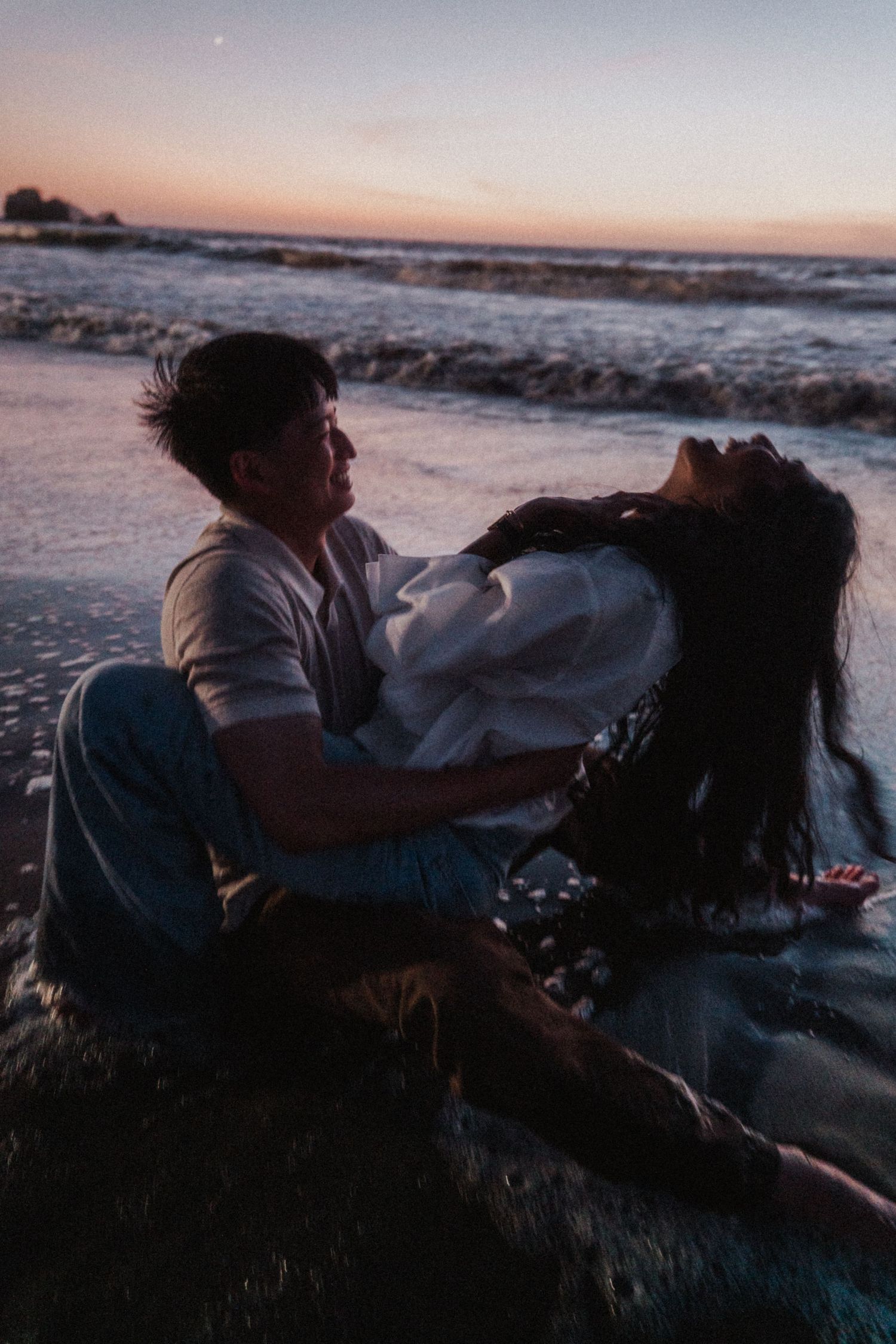 Romantic couple sitting together on a wet beach at dusk with colorful sunset sky behind them.