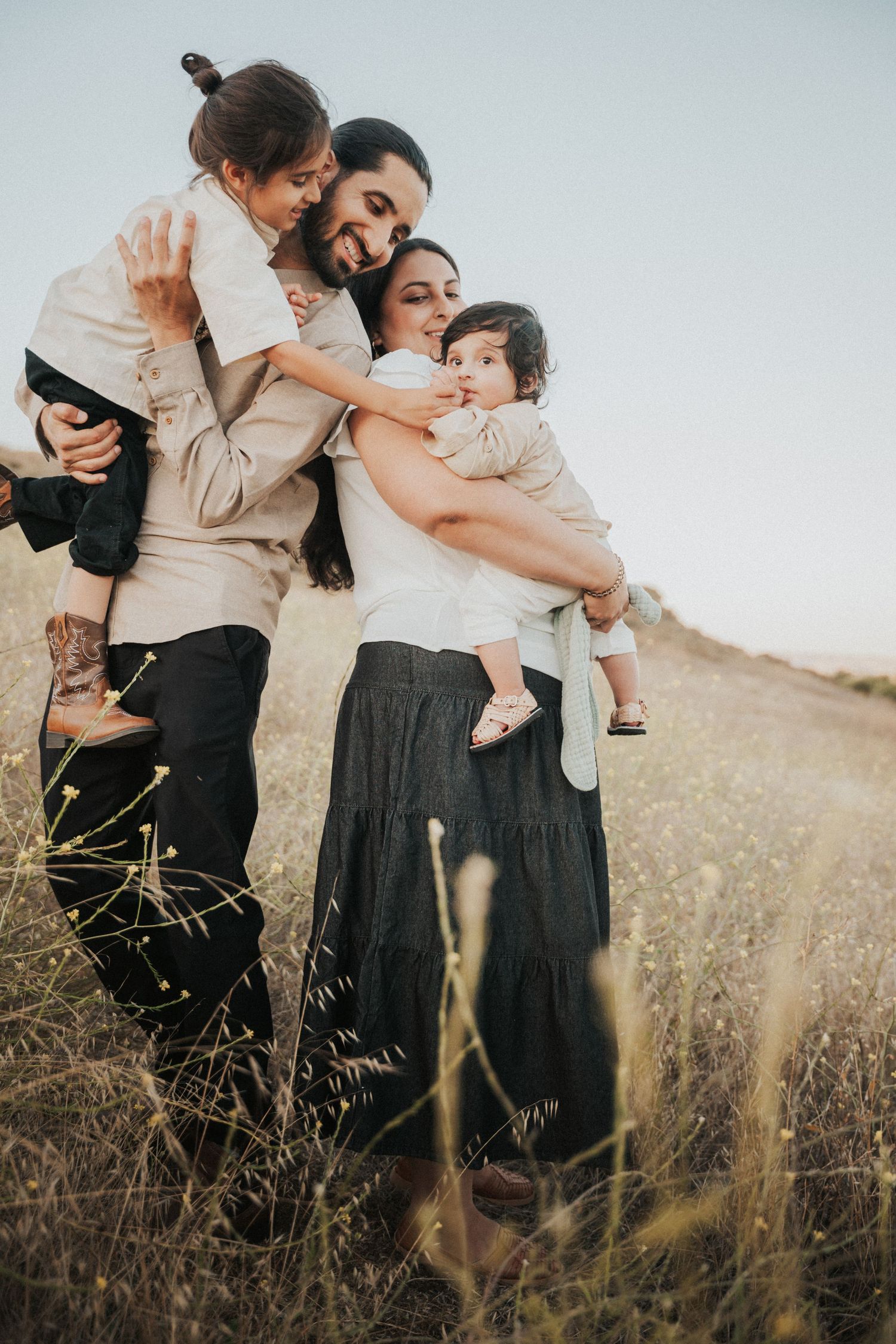 A happy family of four embraces in a golden field, parents holding two young children in warm, natural light. Coyote Hills Regional Park