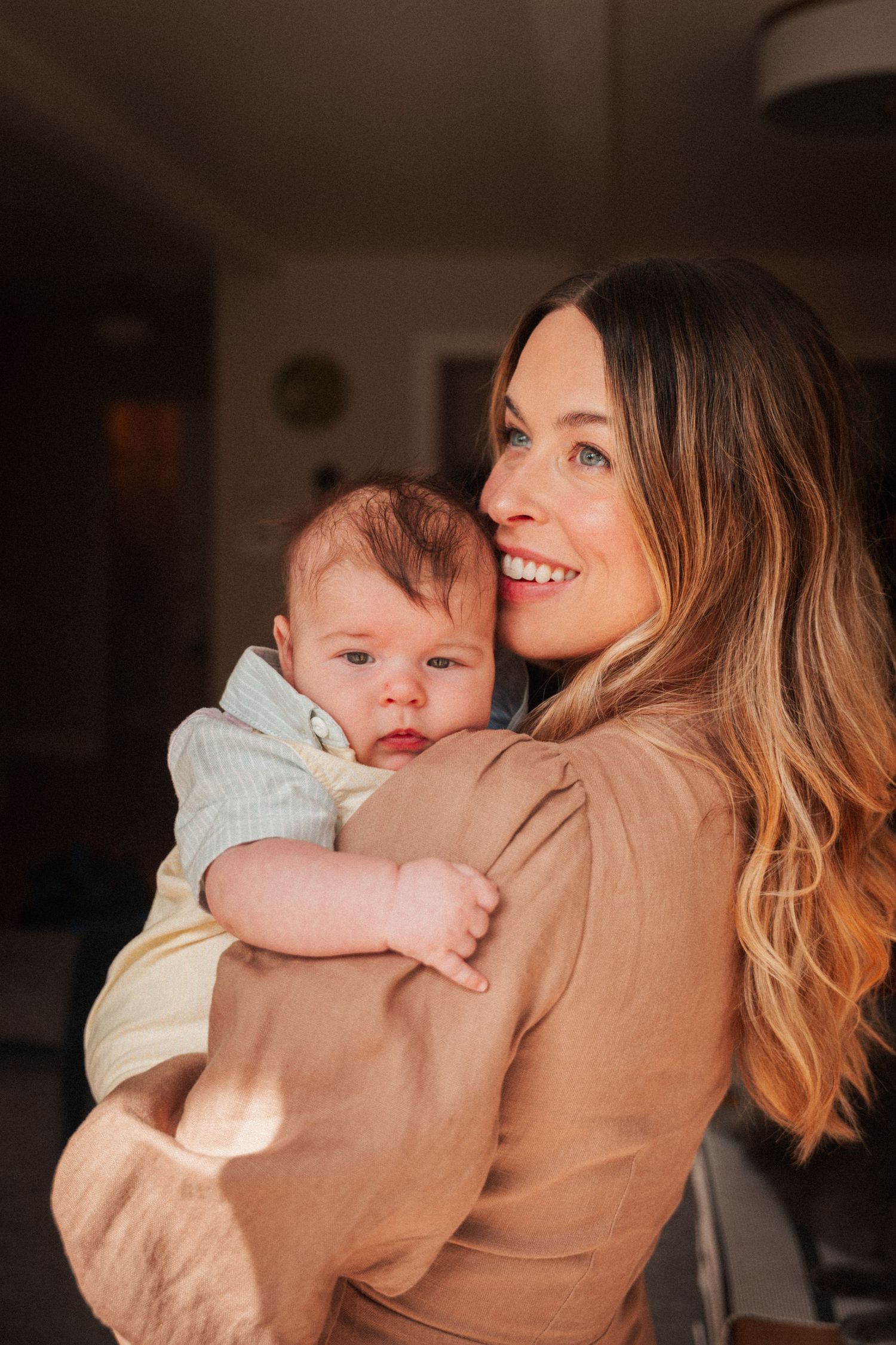 Smiling woman with long hair holding a newborn baby in a warmly lit home setting.