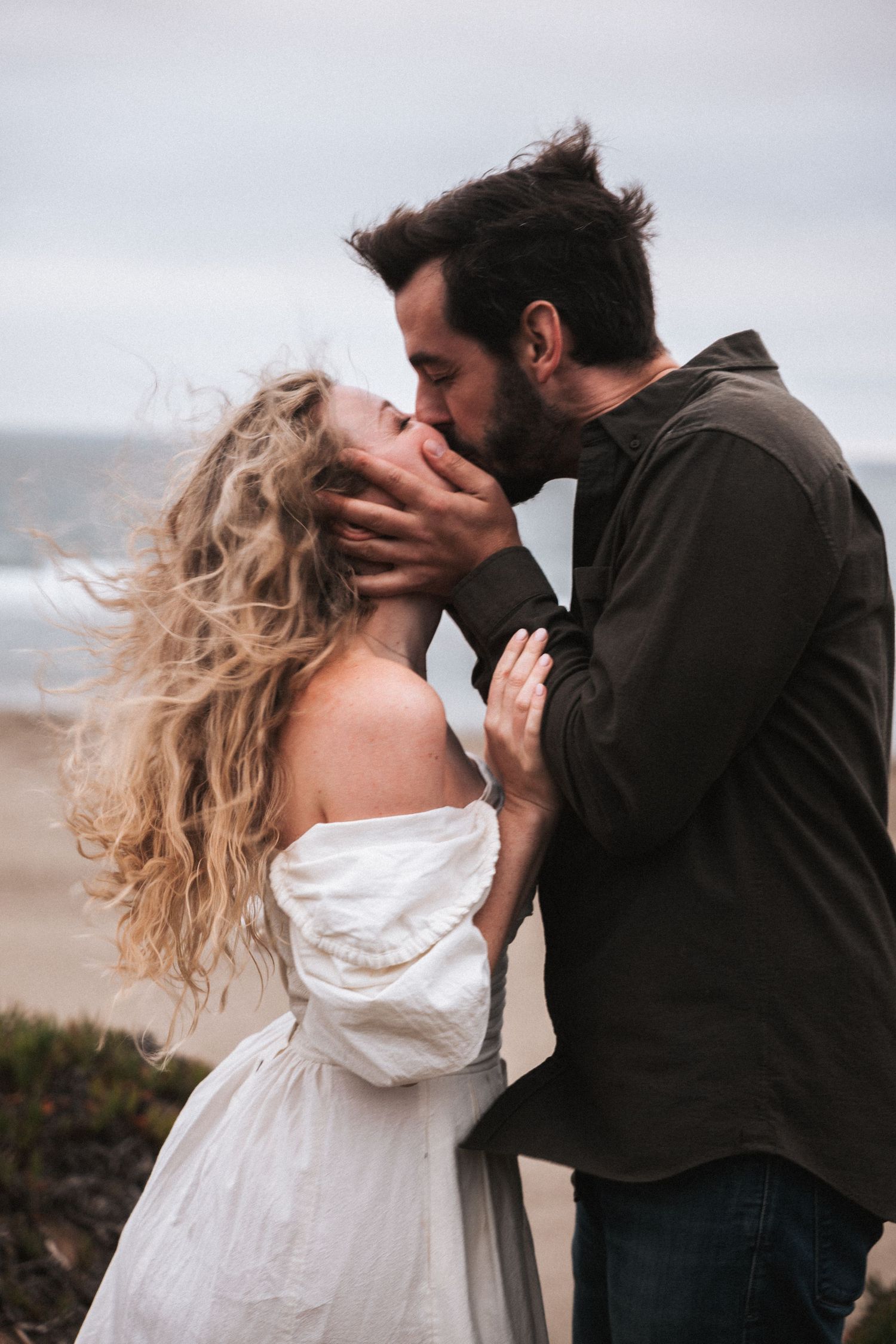 Couple sharing a romantic kiss on a windy beach, woman in white off-shoulder dress, man in dark shirt.