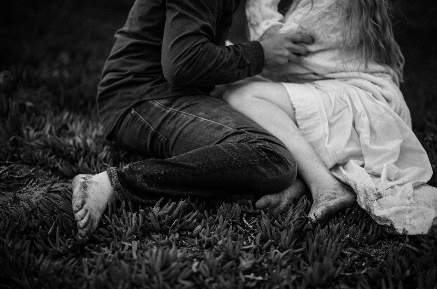 Black and white photo of a couple sitting on grass, bride in white dress and groom in plaid pants.