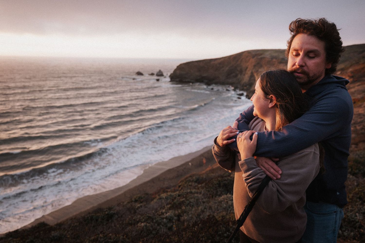 Couple embracing romantically on a coastal cliff at sunset with ocean waves and rocky sea stacks in the background.