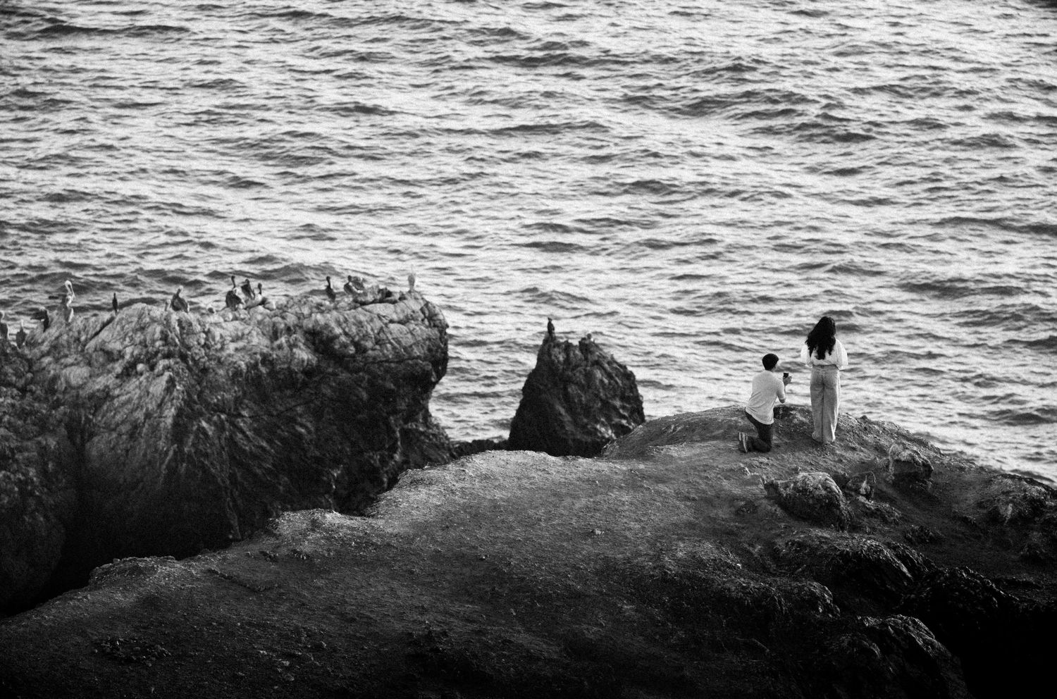 Black and white photo of two people standing on rocky coastal cliffs overlooking the ocean waves.