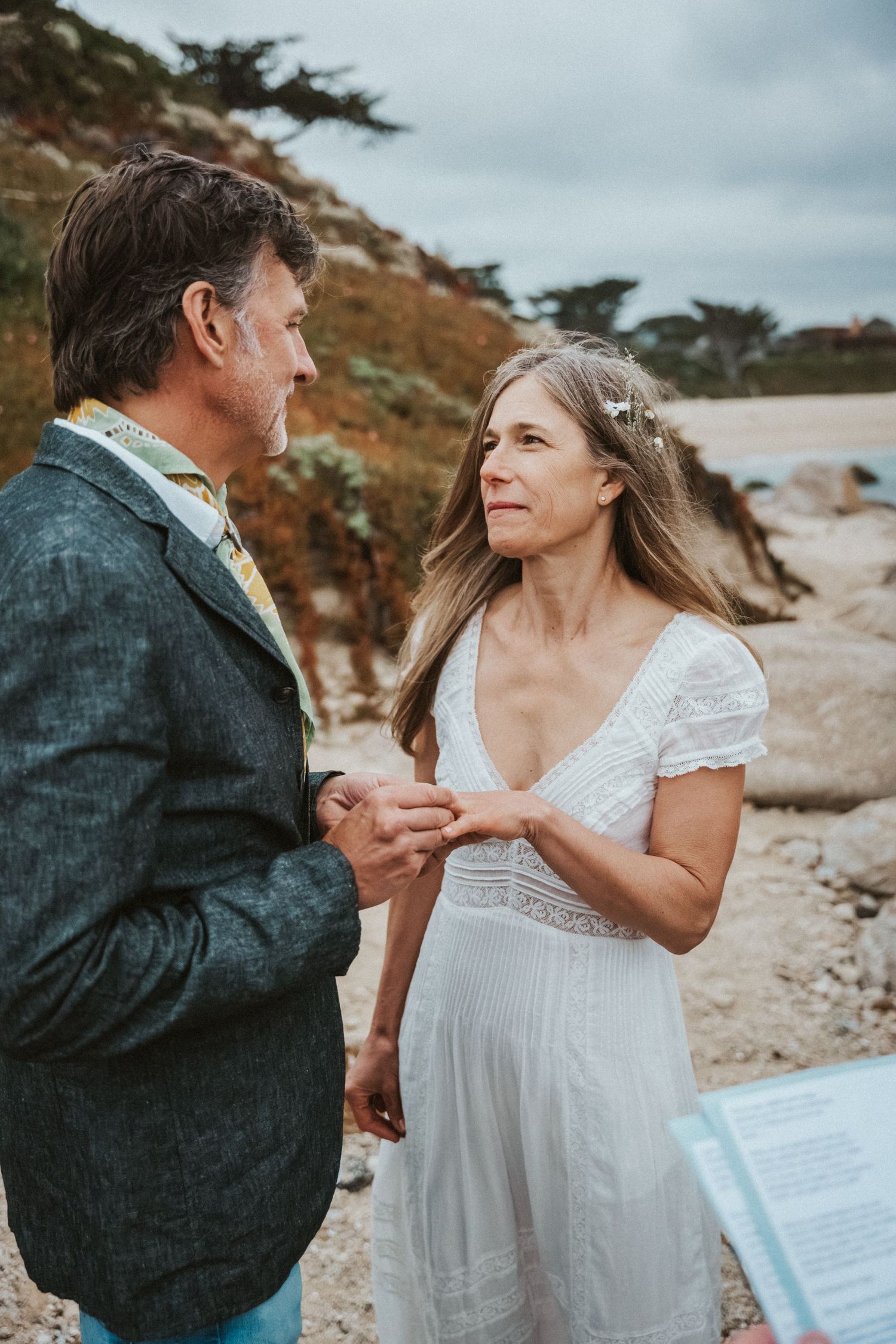 Mature couple exchanging vows in white dress and gray suit at a scenic beach wedding ceremony.