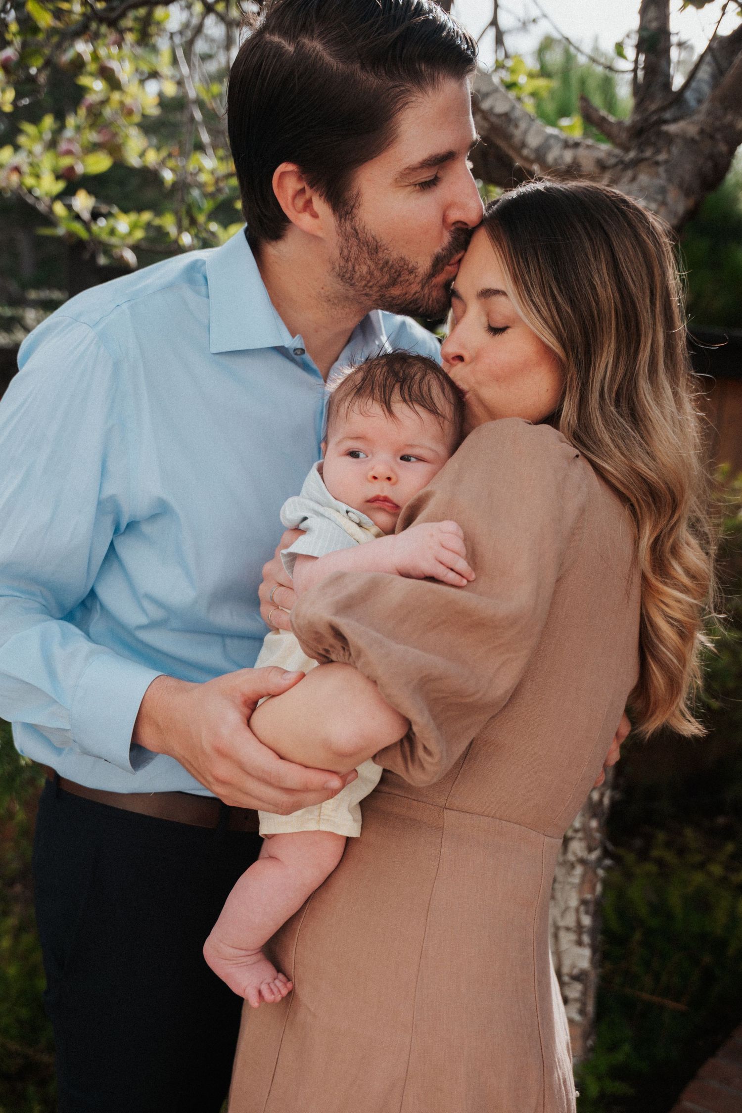 A couple kisses their newborn baby outdoors near trees, man in blue shirt and woman in tan dress.
