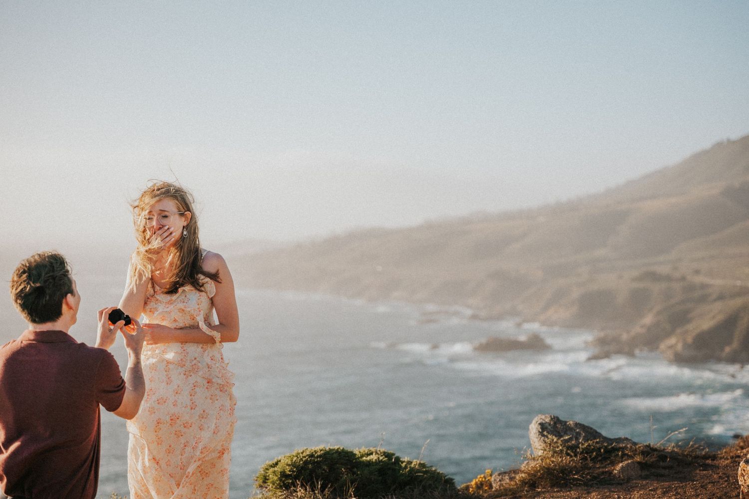 Man proposing to woman on coastal cliffside at sunset with ocean and mountains in background.