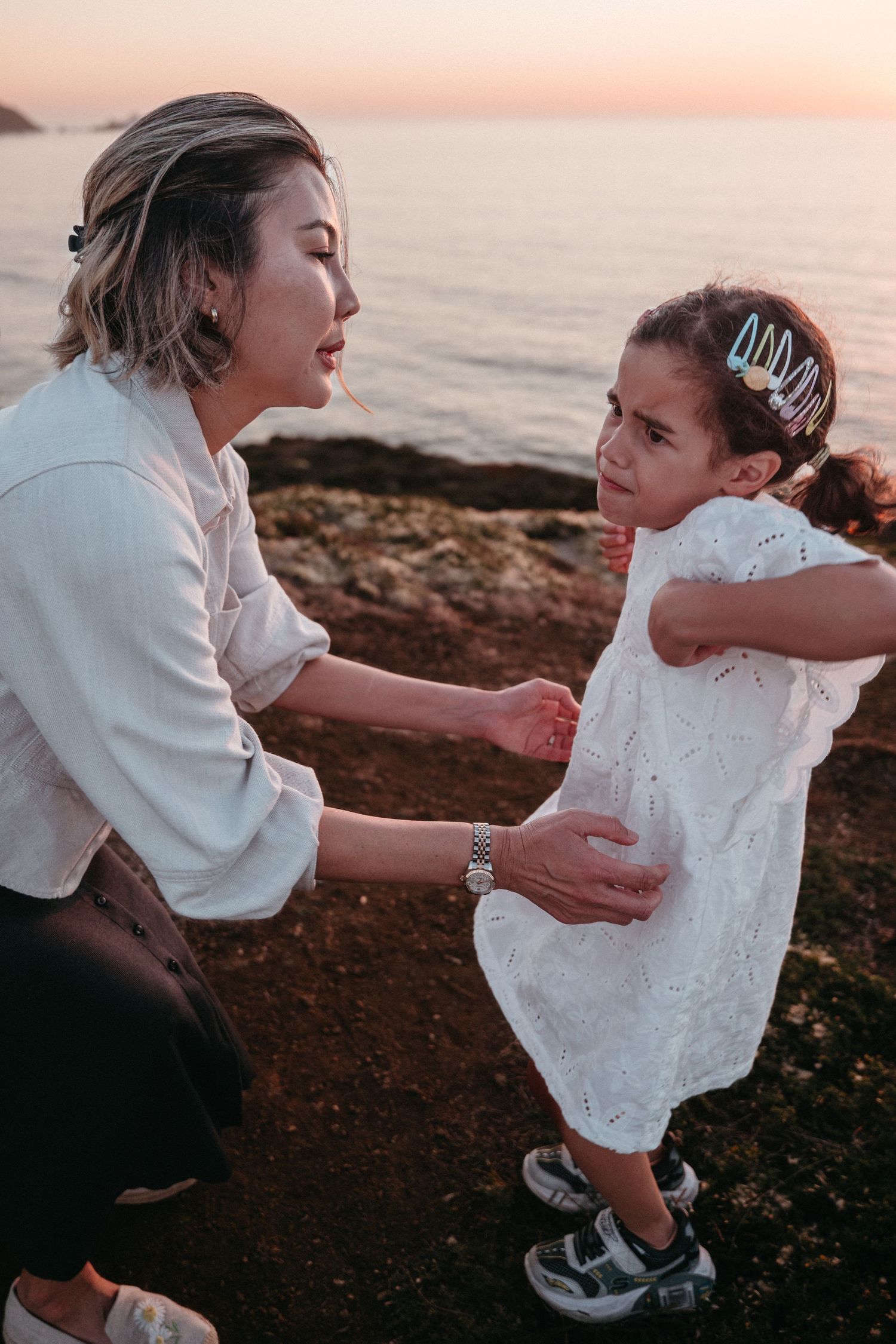 A mother crouches down to speak with her young daughter in a white dress near a rocky ocean shore at dusk.
