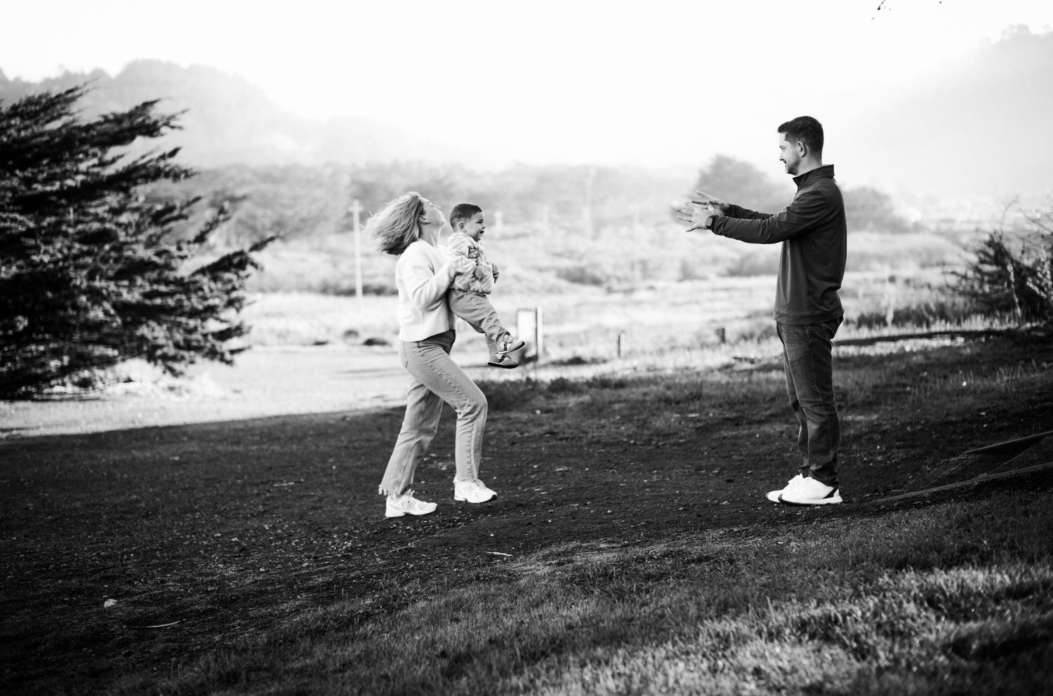 Black and white photo of a couple embracing outdoors while a man photographs them on a scenic hillside.