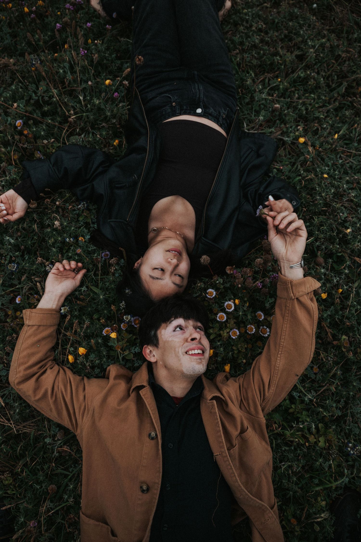 Couple lying on flower-covered grass, smiling and holding hands, wearing casual jackets in an outdoor setting.