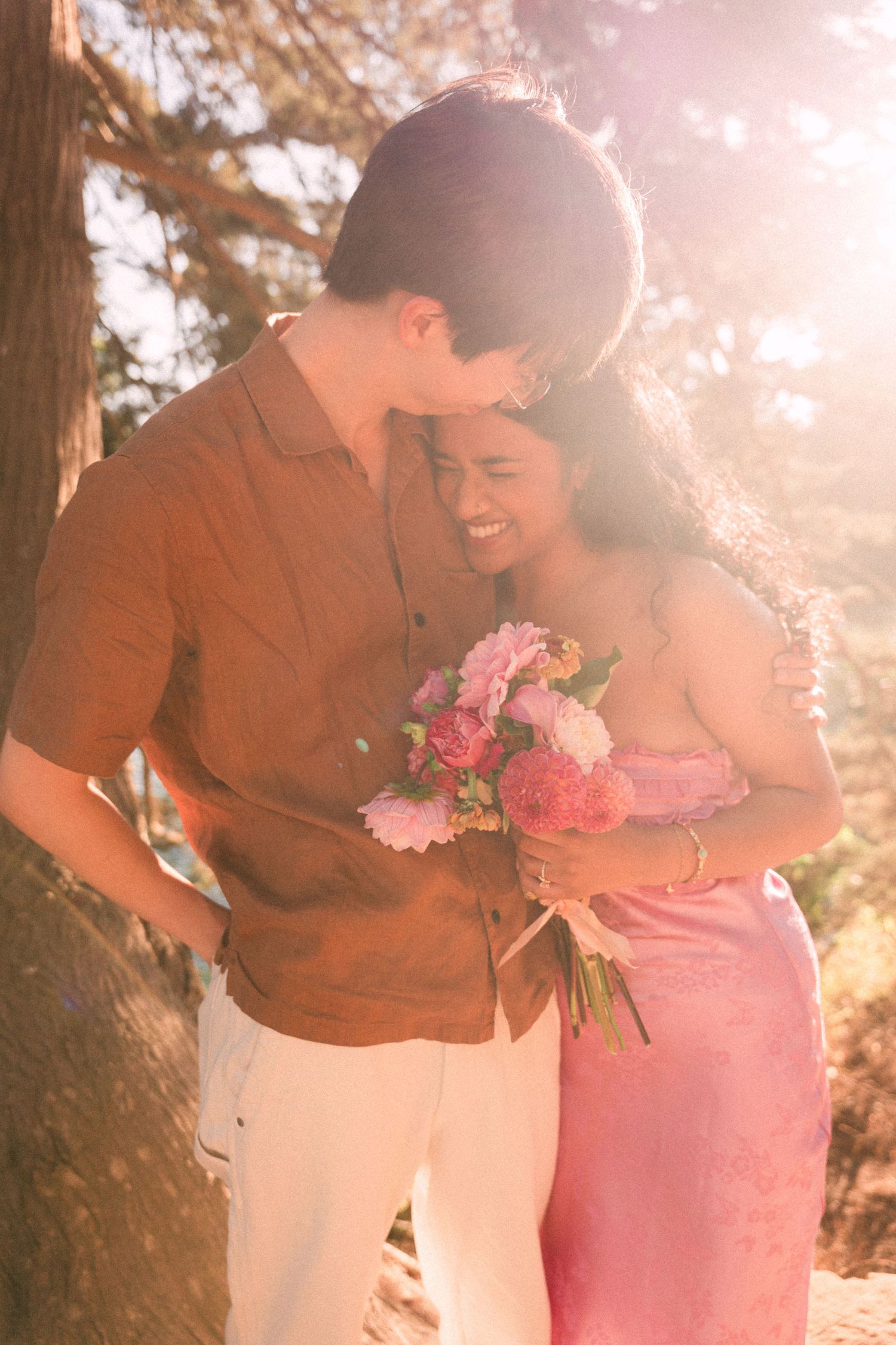 A couple shares a romantic moment outdoors, woman holding pink flowers, bathed in warm golden sunlight near a tree.