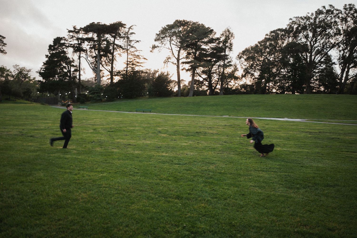Two people playing with a dog on a lush green park lawn at dusk, surrounded by tall trees.