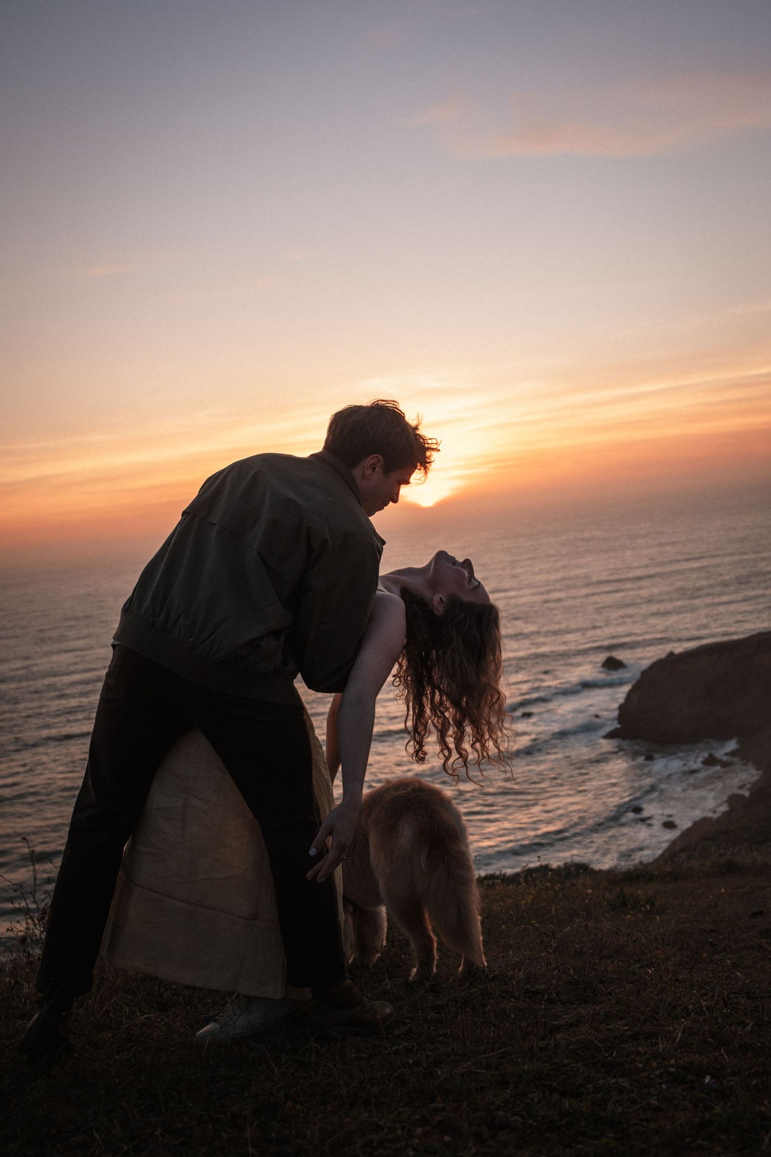 Romantic couple kissing at sunset on a coastal cliff with a dog nearby. Photography by The Y.K. Studio.