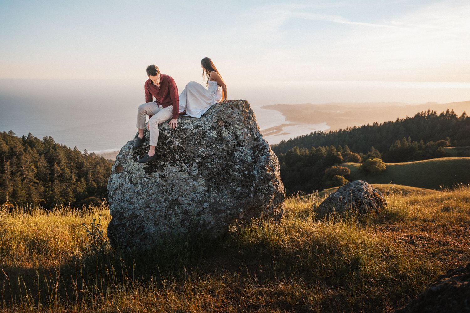 Couple sitting on large boulder at sunset overlooking scenic coastal landscape with forest and ocean views.