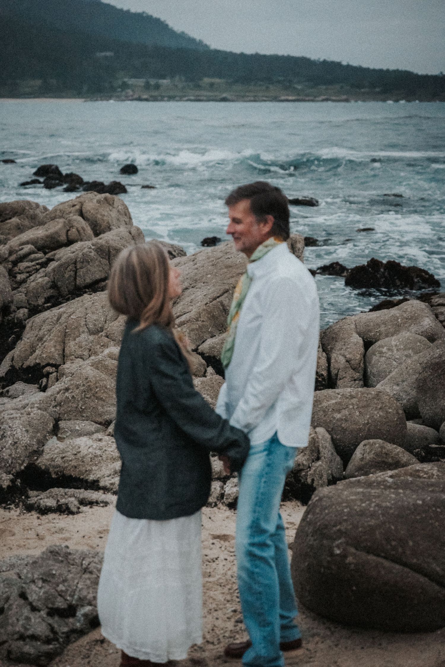 Couple holding hands on rocky Pacific coast beach, woman in white dress and gray jacket, man in white shirt and jeans.Photography by The Y.K