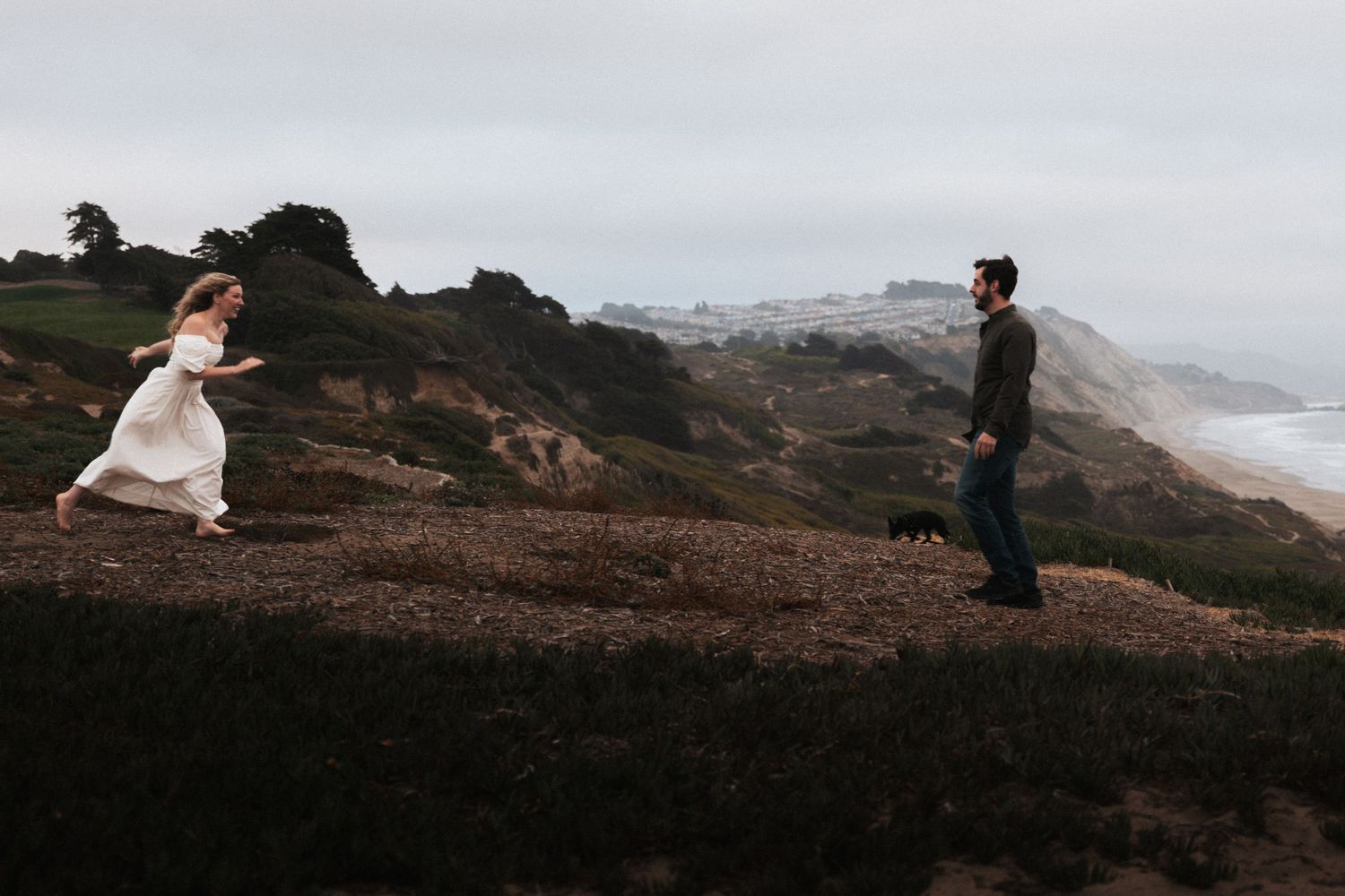 Couple in wedding attire on coastal cliffside, bride running toward groom with ocean backdrop on overcast day.