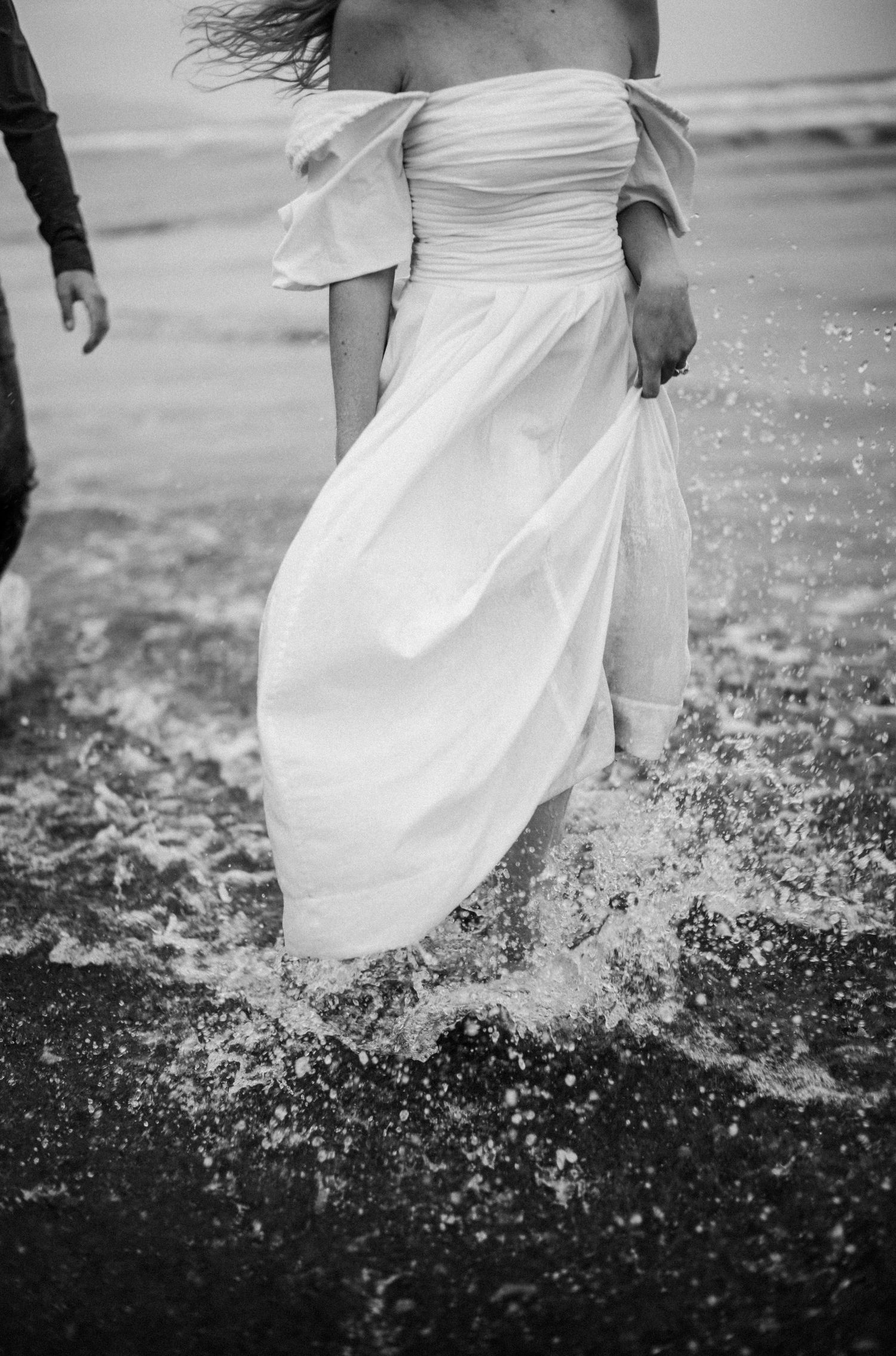 Black and white close-up of a bride's white off-shoulder gown splashing through ocean waves.