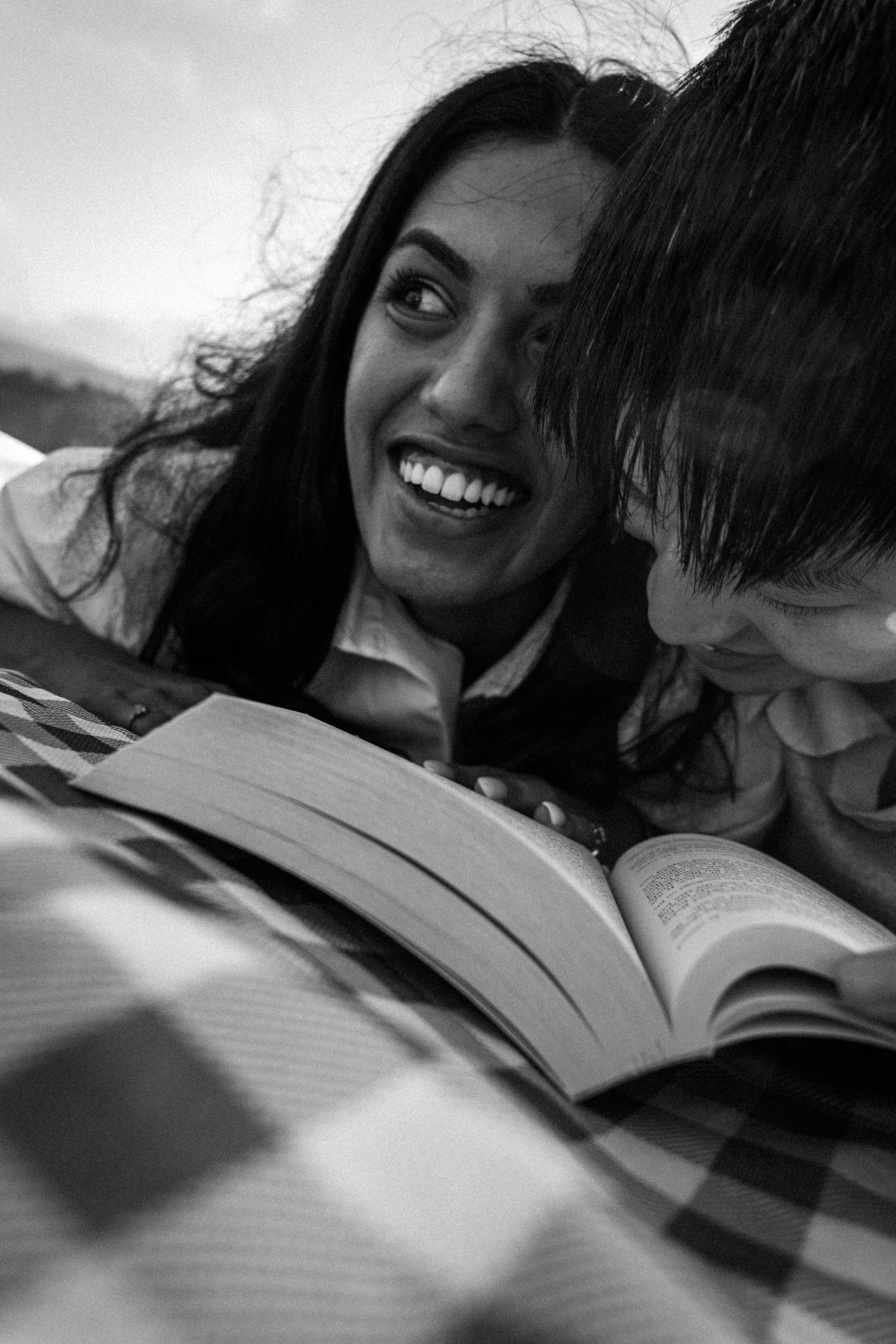 Black and white photo of a smiling woman and man lying down reading a book together on a checkered blanket.