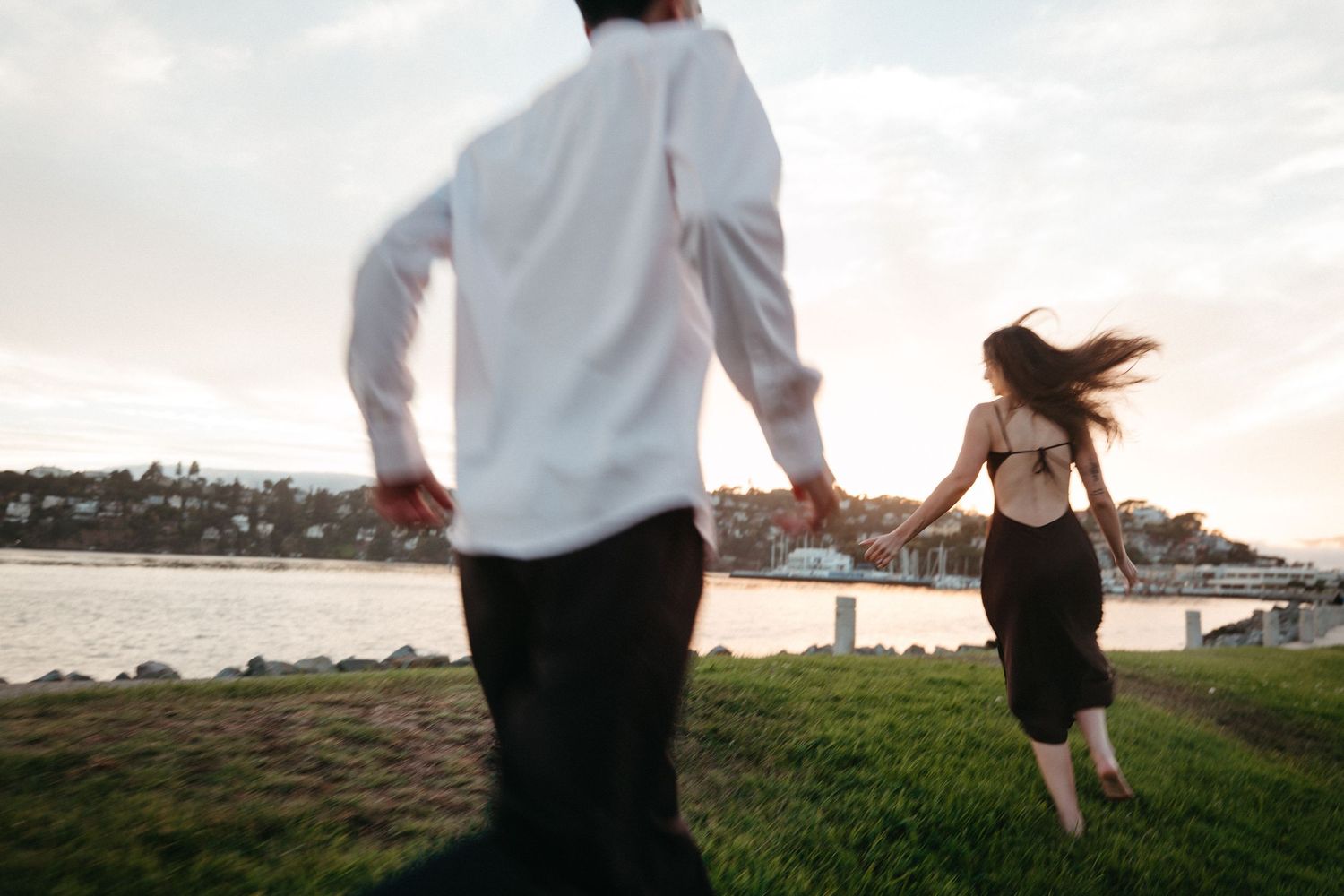 Couple holding hands running on grass by waterfront at sunset, woman in black dress with open back, man in white shirt.