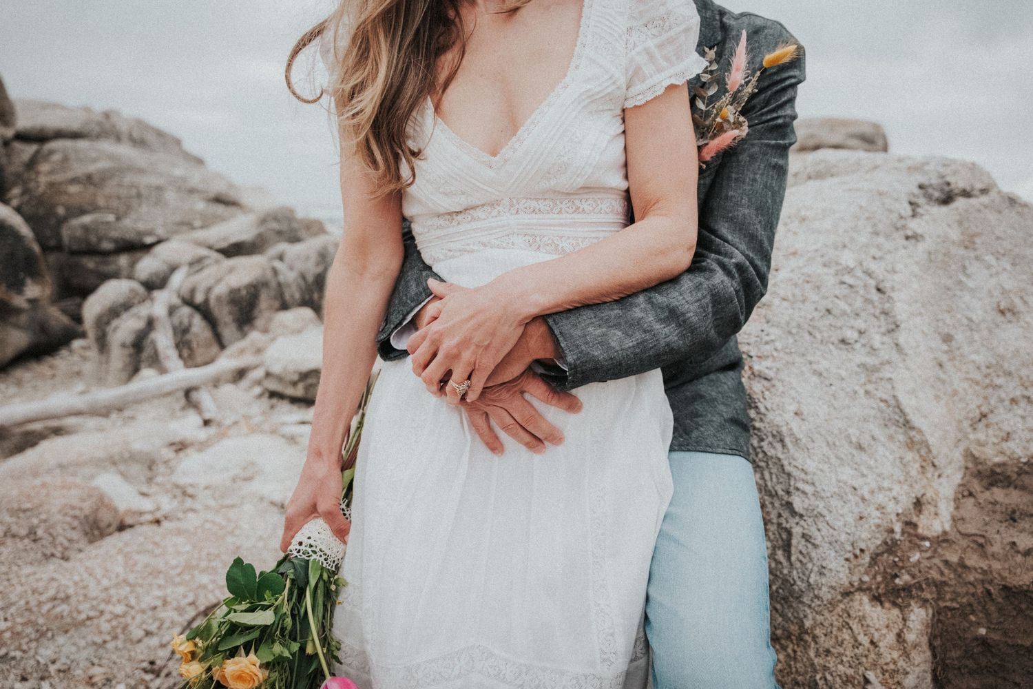 Bride in lace wedding dress embraced by groom holding yellow bouquet on rocky beach setting.