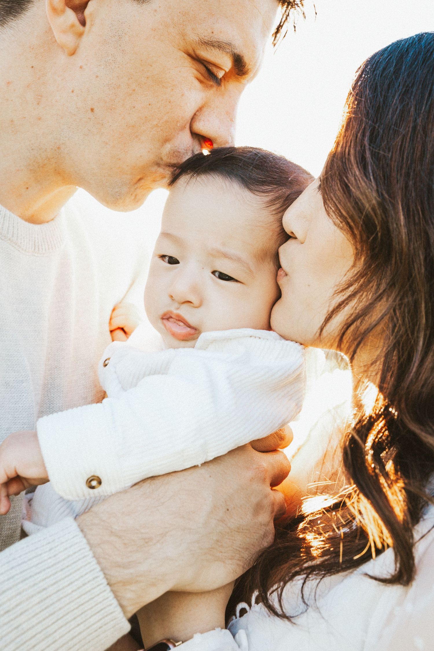 Parents kissing their baby on both cheeks in a warm golden backlit family portrait.