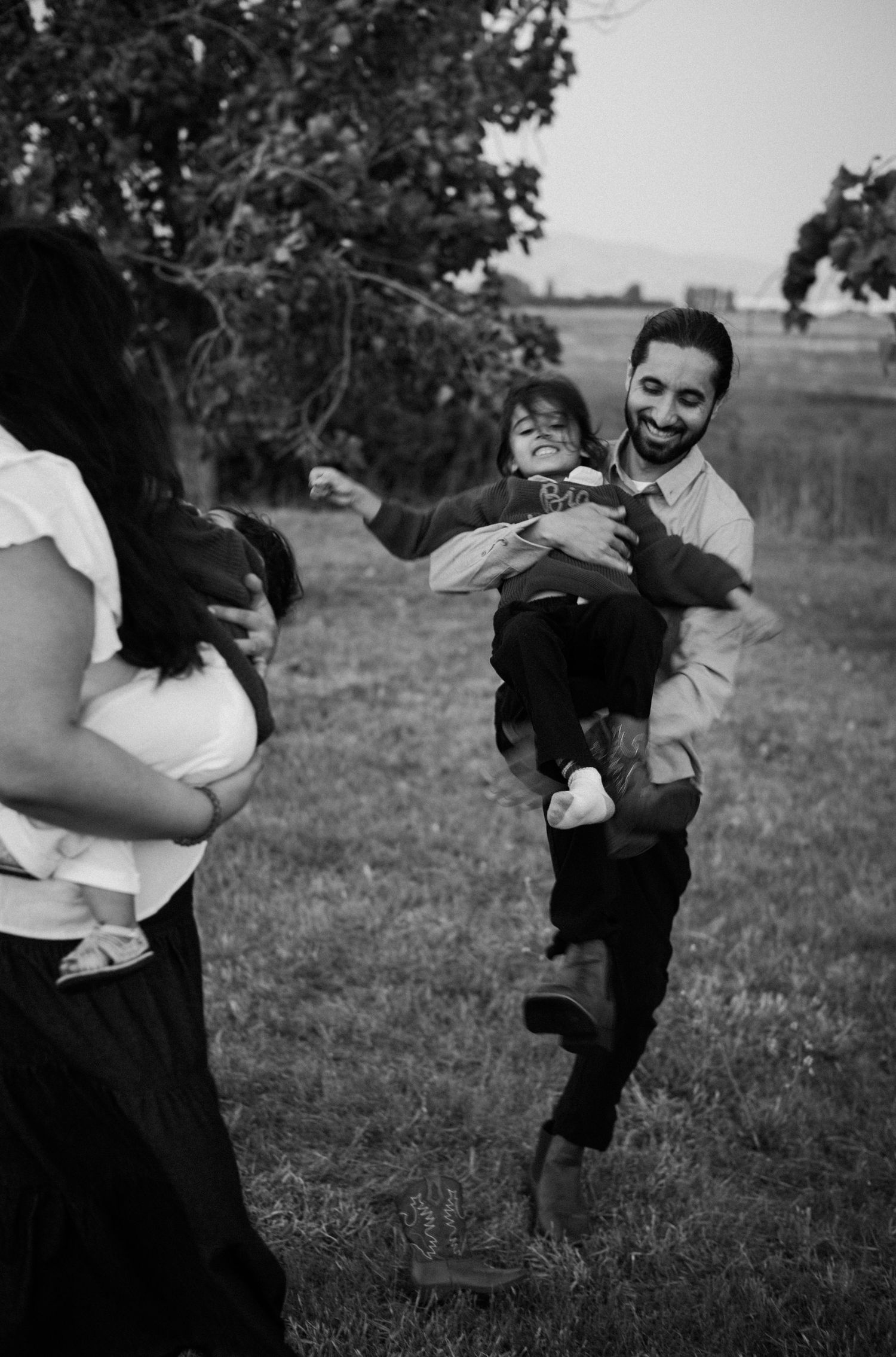 Black and white photo of a laughing father lifting his child while mother watches in a grassy outdoor setting.
