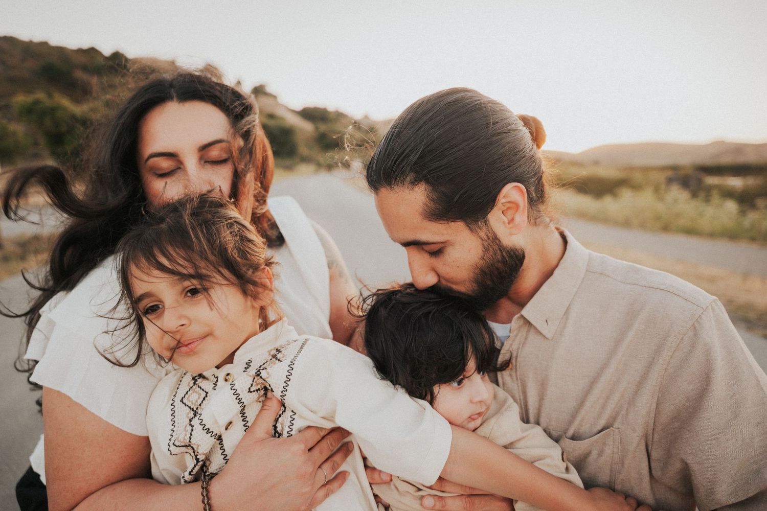 A loving family of four embraces outdoors at sunset, parents cuddling two young children in a warm, tender moment.