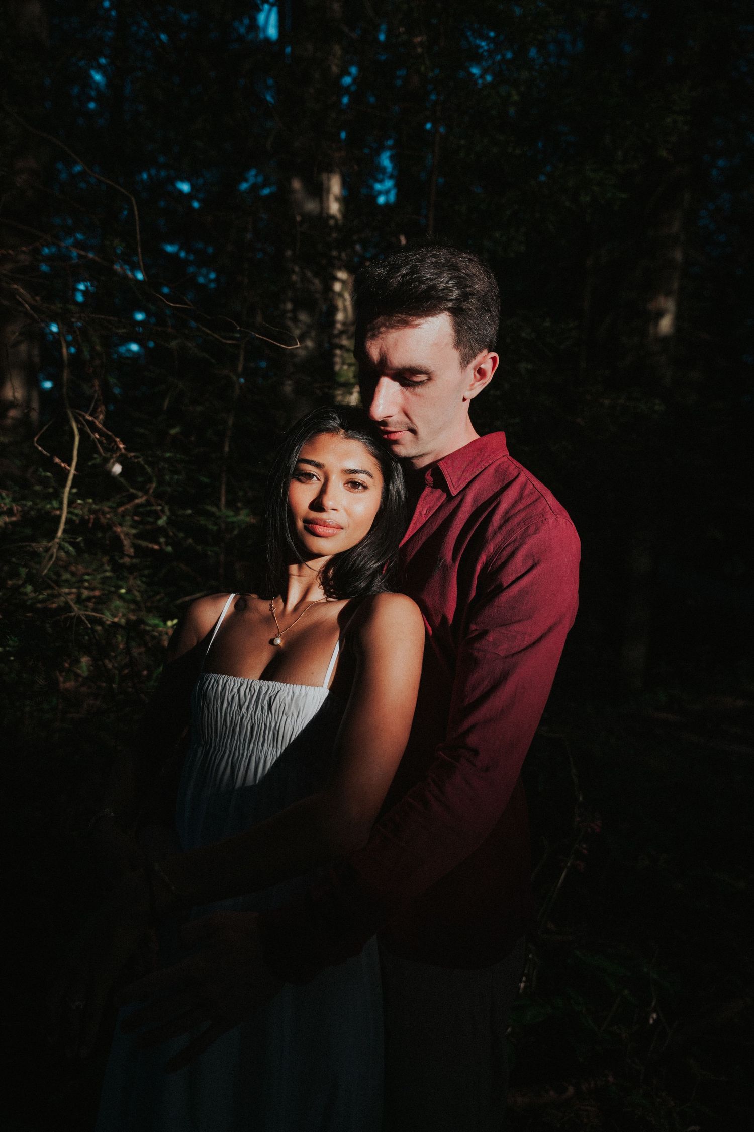 Couple embracing in a dark forest, woman in white dress, man in red shirt, dramatic moody lighting.