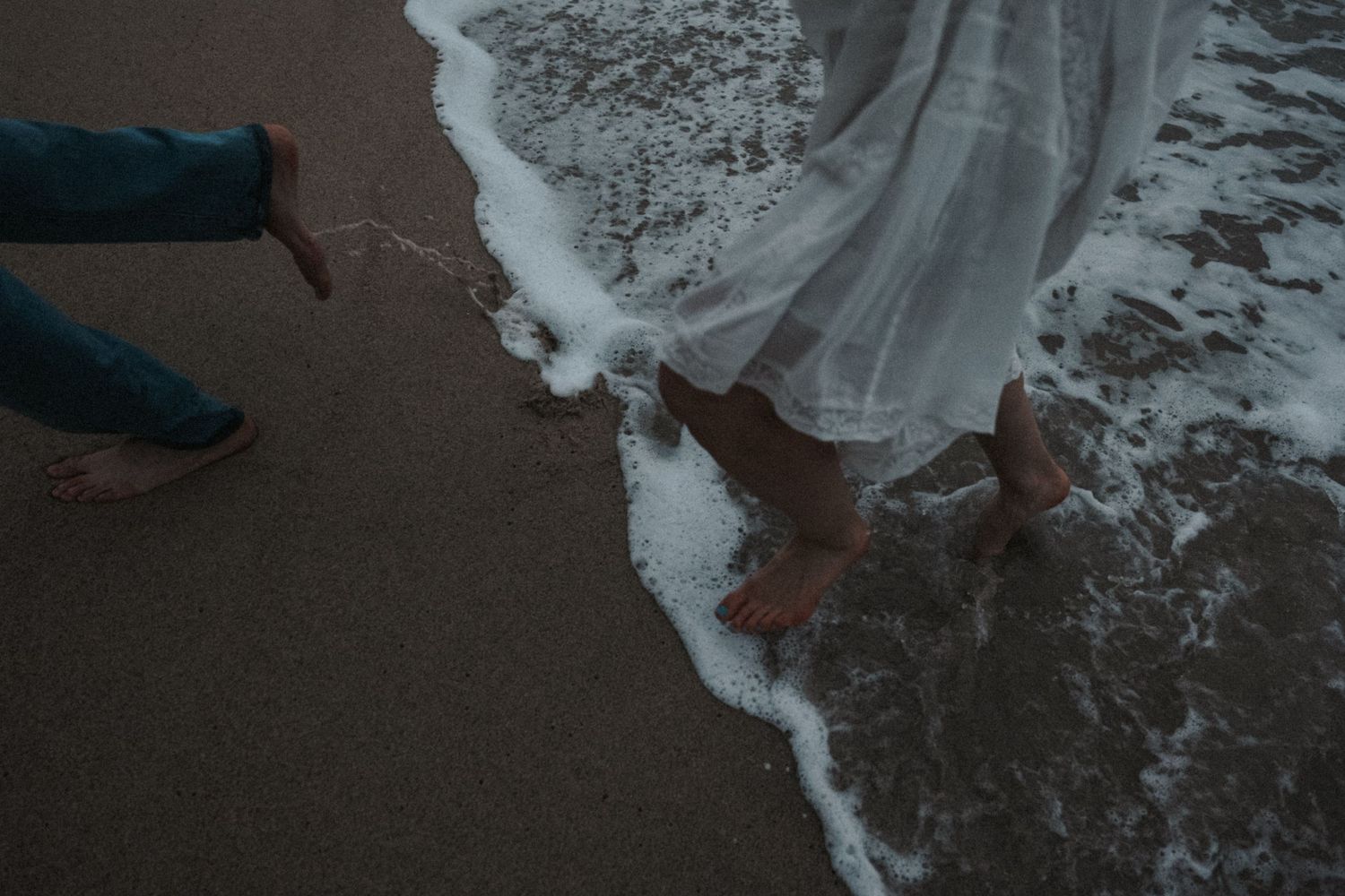 Barefoot couple standing at ocean shoreline with white wedding dress hem touching the waves.