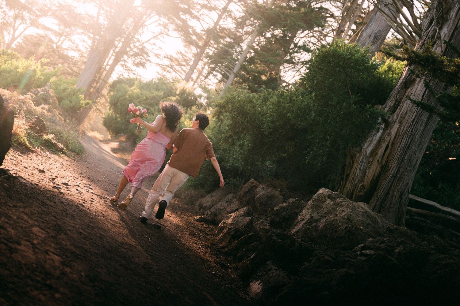 Two children run hand-in-hand up a sunlit forest trail surrounded by tall trees and rocky terrain.