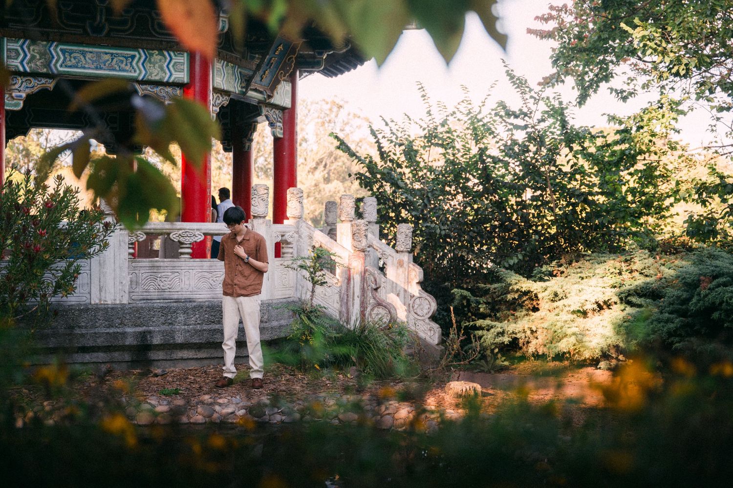 Man standing in a lush garden near a traditional Chinese pavilion with red columns and ornate architecture.