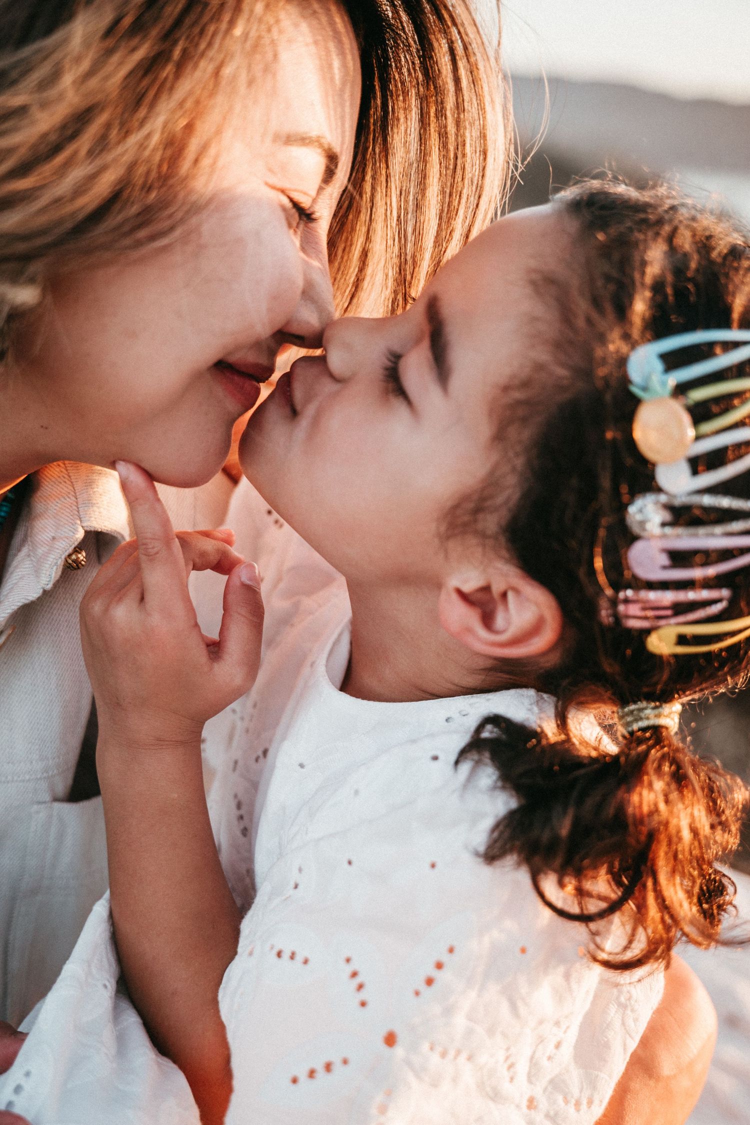 A mother and young daughter share a gentle nose-to-nose kiss in warm sunset light.
