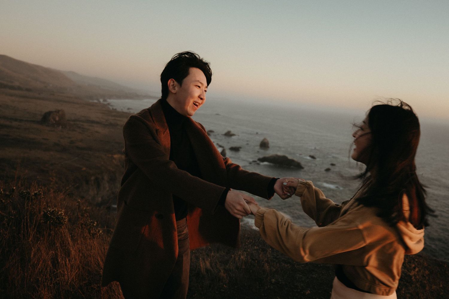 A smiling couple holds hands and dance on a grassy coastal hillside overlooking the ocean at sunset.