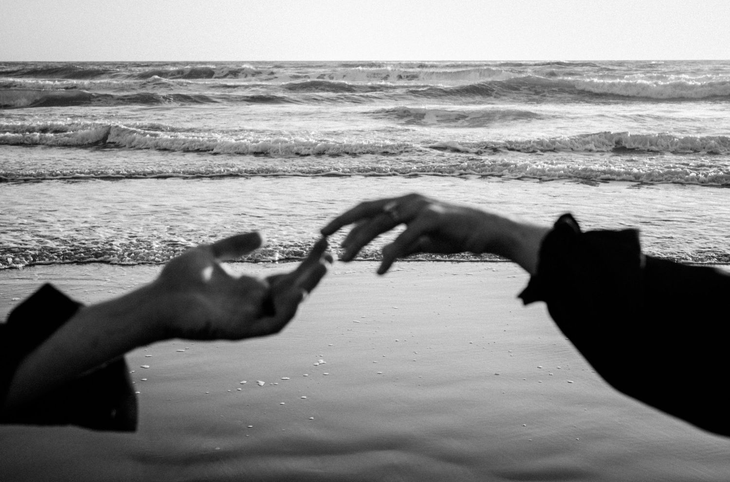 Two hands reaching toward each other on a black and white beach with ocean waves in the background.