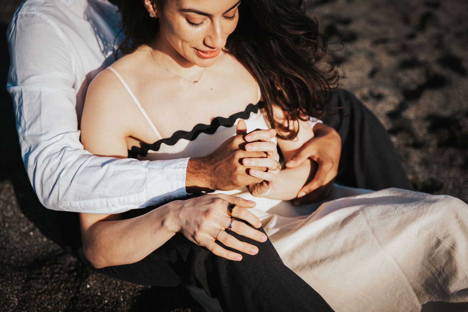 Couple sitting closely on beach, man embracing woman from behind, golden sunset light.
