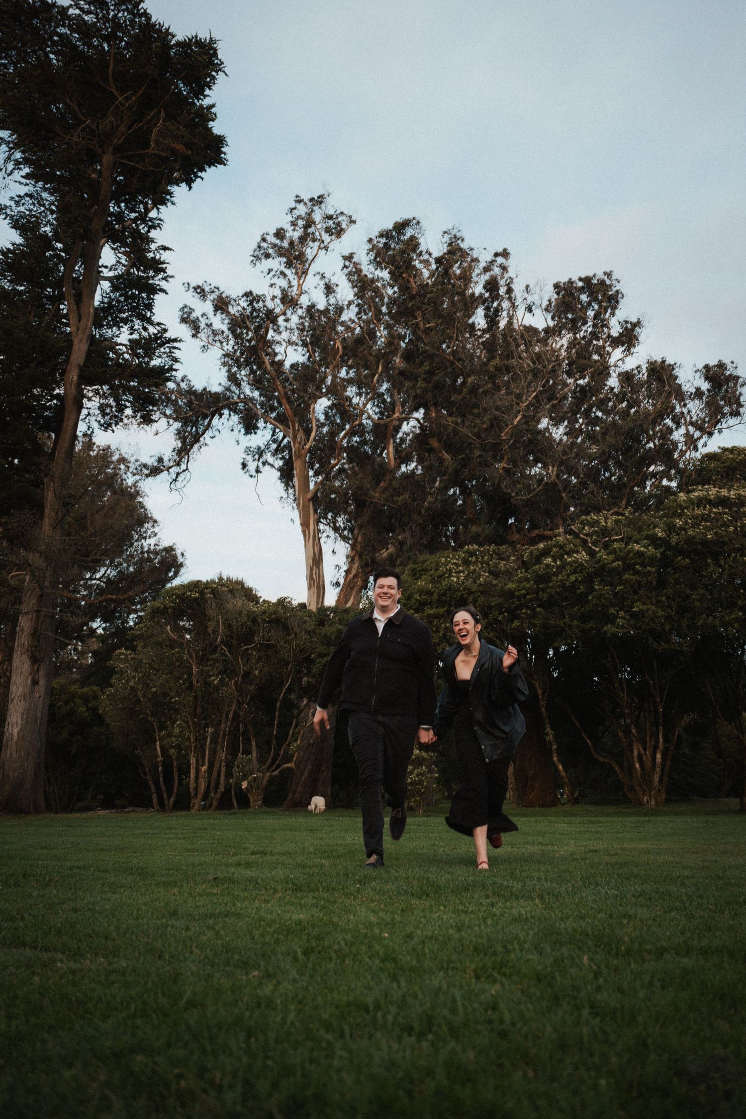Joyful couple in black outfits running hand in hand across a green lawn with tall trees behind.