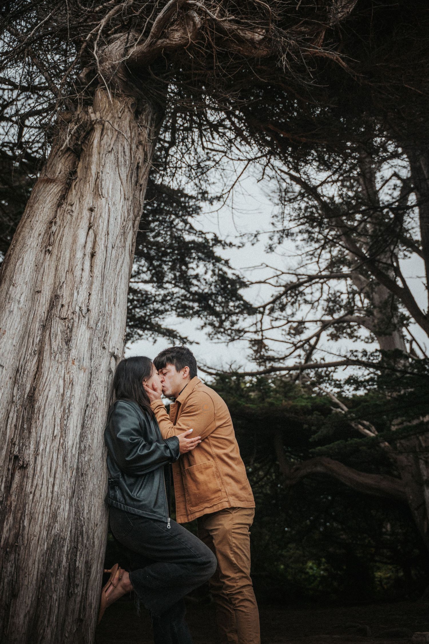 Romantic couple sharing a kiss leaning against a large tree trunk in a moody outdoor setting.