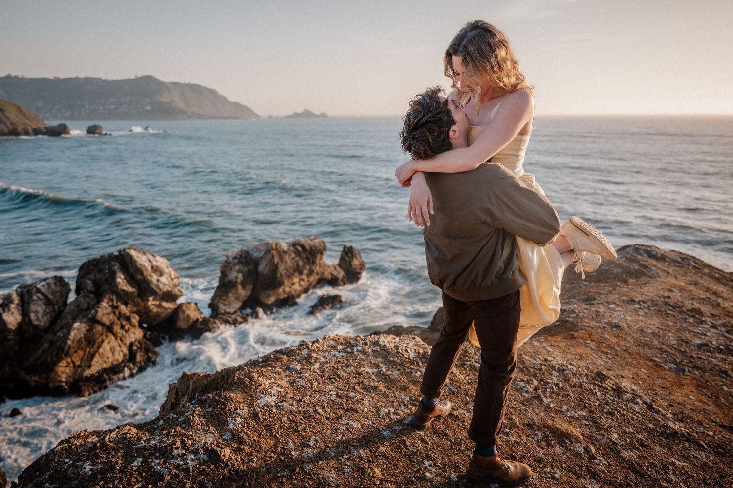 Couple embracing romantically on rocky coastal cliffs at sunset with ocean waves and mountains in background.