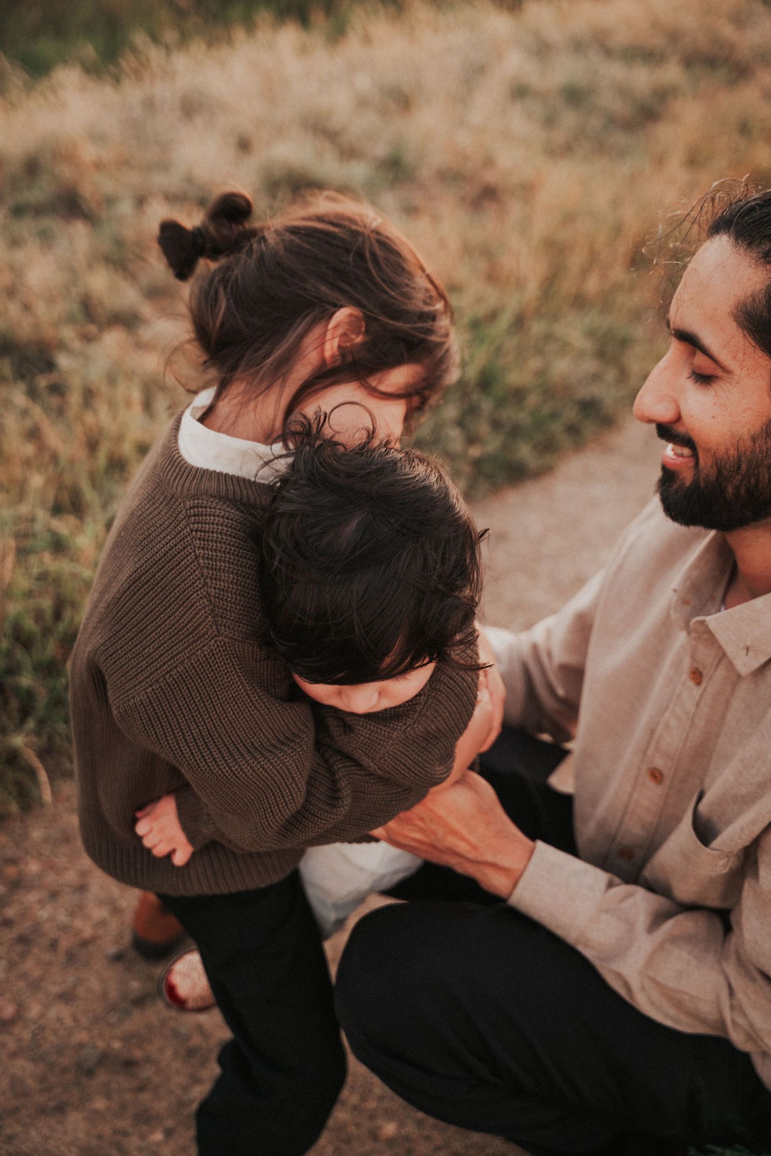Bearded father smiling as young daughter hugs baby sibling on a grassy trail.