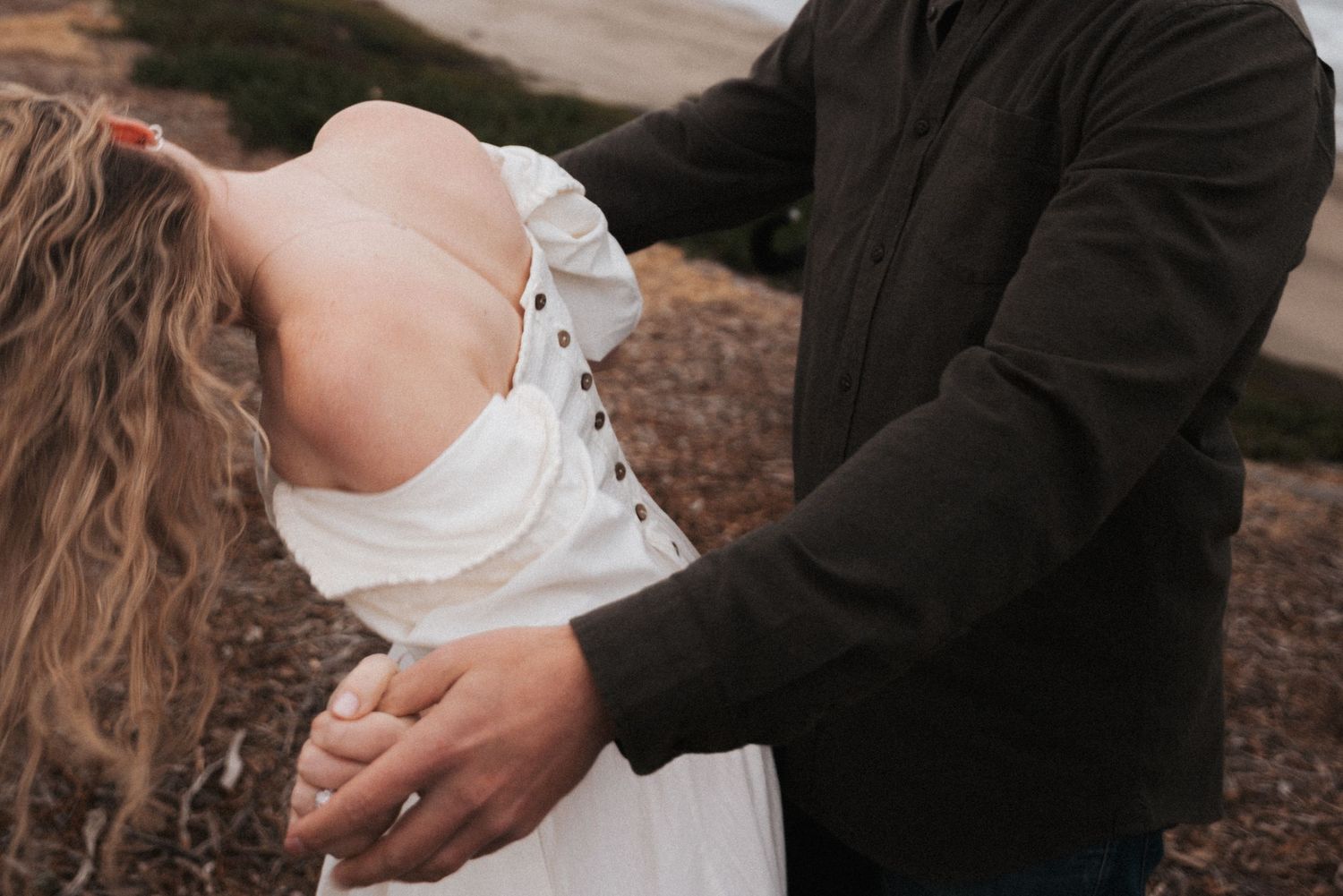 Couple dancing outdoors, woman in white off-shoulder dress with curly hair, man in dark jacket holding her hands.
