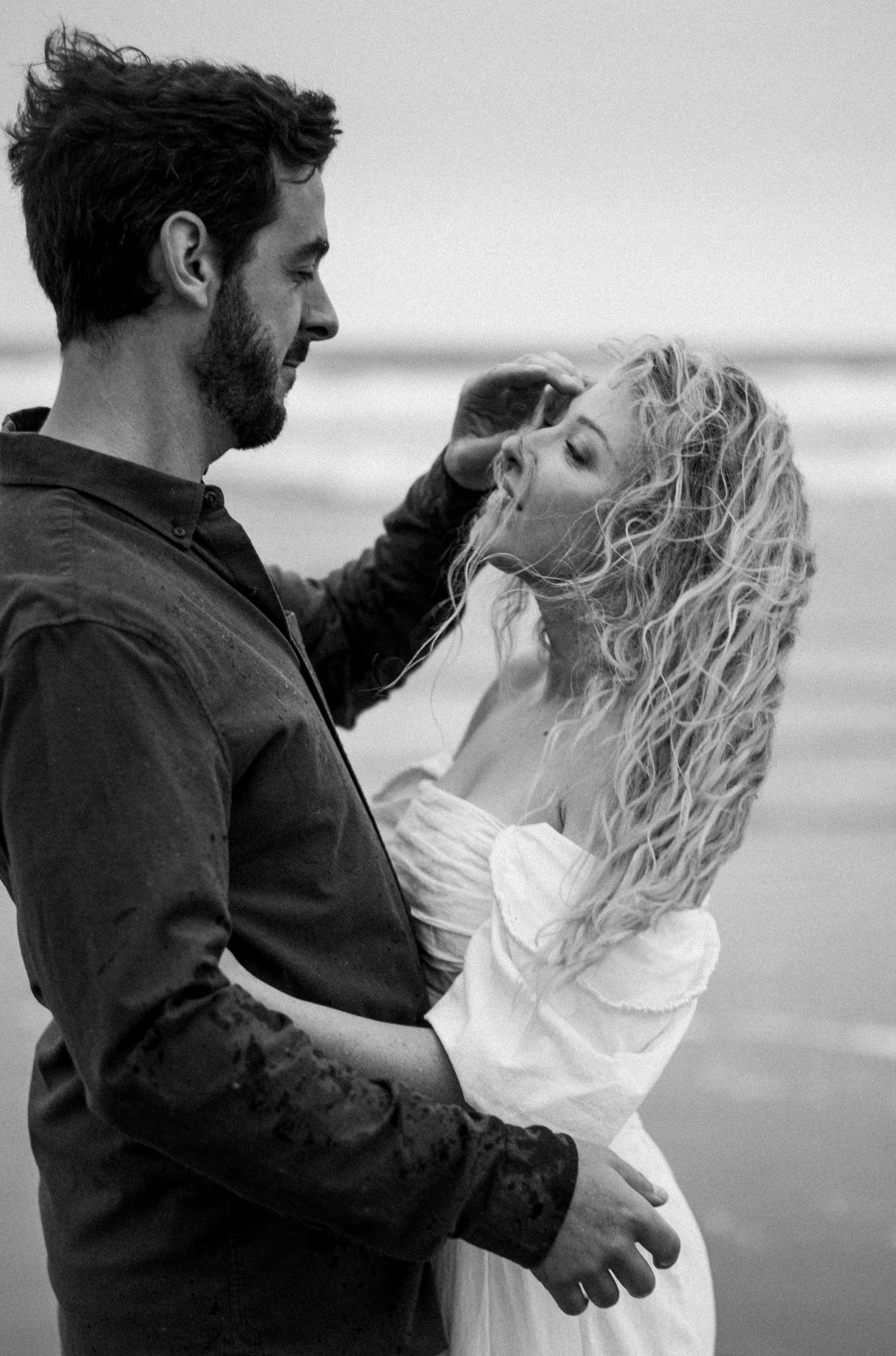 Black and white photo of couple embracing on beach, man brushing curly hair from womans face in white dress.